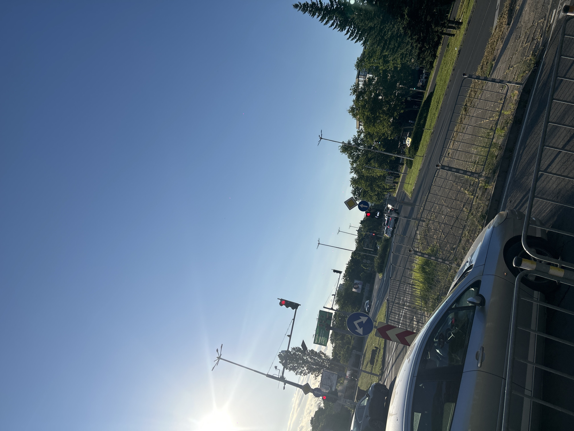 The image depicts an outdoor scene with a clear blue sky, suggesting a sunny day. The perspective is from inside a vehicle looking outwards and slightly upwards. A silver car is prominently visible in the foreground, taking up the right side of the frame. It appears to be parked or moving slowly behind a metal barrier.
Further down the road, traffic lights are visible, with one displaying a red light, indicating a stop. Several road signs are also present, including a diamond-shaped yellow sign with a black border, and a circular blue sign with white arrows indicating a mandatory turn to the left. A red and white chevron sign is also visible on the right side of the road.
The setting appears to be a city street or highway, with a paved road, a grassy verge, and trees lining the side. In the background, buildings can be seen amongst the trees, and more traffic is visible further down the road, including what appears to be a yellow taxi. A tall street light with multiple arms is also visible.
The sun is bright and low in the sky, casting a lens flare towards the left side of the image, suggesting it is either early morning or late afternoon. The overall atmosphere is that of a typical day in an urban or suburban environment with traffic activity. No people are clearly discernible in the image. There is no visible text that is legible.