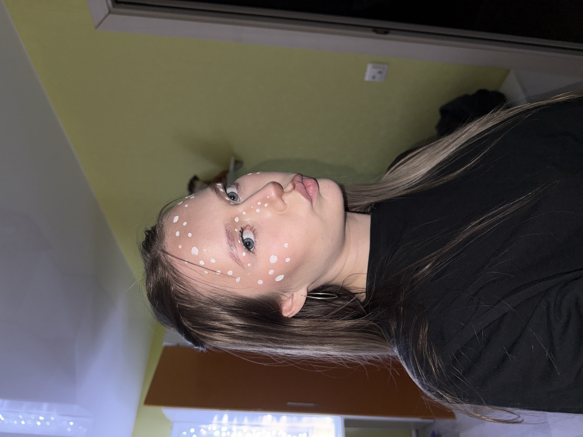A young woman with light skin and blue eyes is positioned in the center of the image. She has brown hair with blonde highlights. The woman's face is adorned with white dots. She wears a silver hoop earring in her left ear and a black top. The background shows a light green wall near a window frame, a beige wall, and a string of lights. The location is not specified as Zhigalki, Russia.