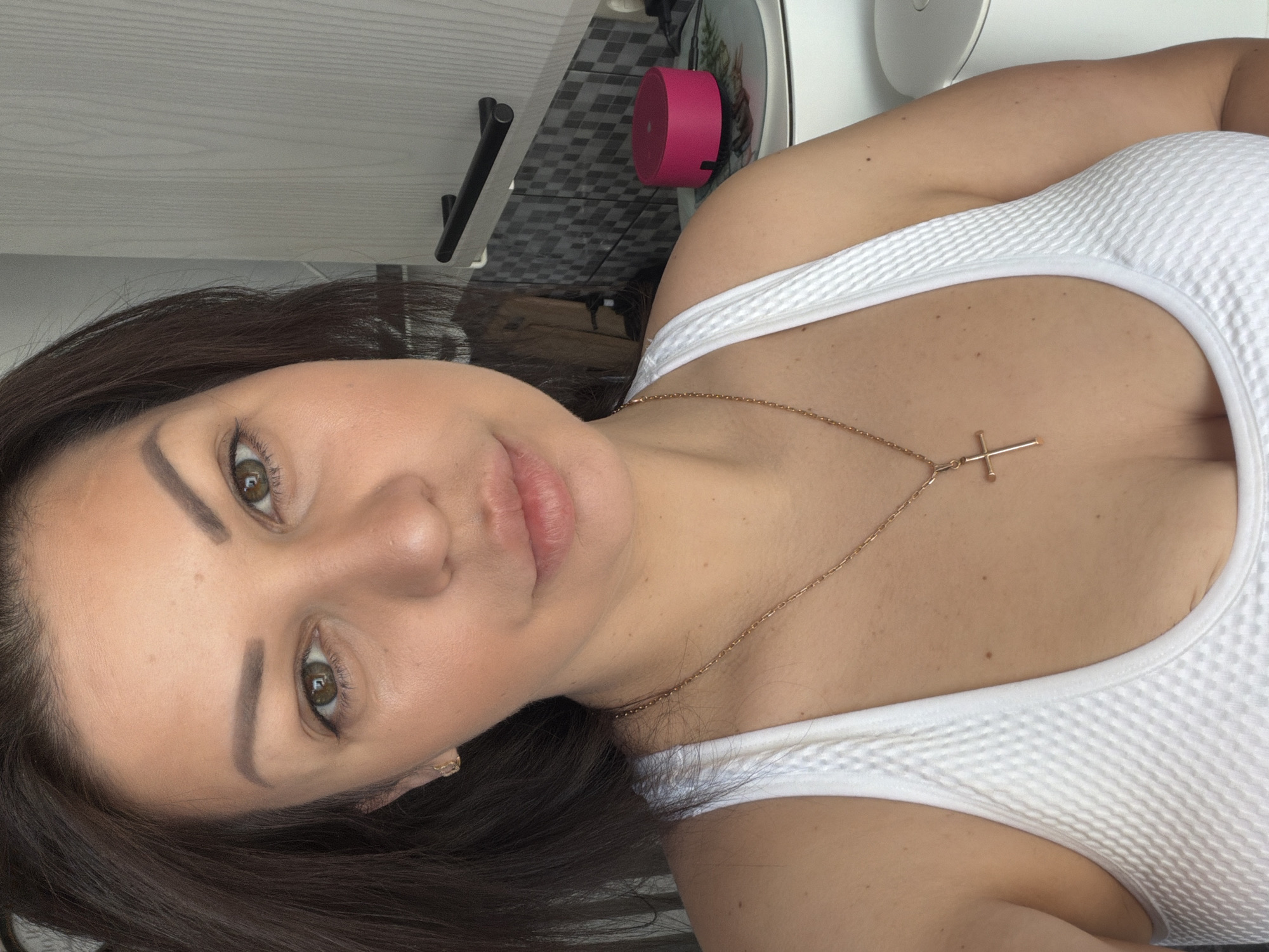 A selfie depicts a woman, likely in her 20s or 30s, inside a residence in the city of Sochi, Russia. She possesses medium-dark brown hair, green eyes, and an olive skin tone, with well-defined eyebrows. Her expression is neutral as she directly faces the camera. She is wearing a white textured tank top and a gold chain necklace with a plain gold cross pendant. A small gold hoop earring is visible in her left ear. The background behind her left shoulder features a light-colored wall or cabinet. To her right, a section of grey square tiles arranged in a checkerboard pattern is visible, above which a black horizontal handle is mounted. On a surface in the background, a bright pink cylindrical object, a clear glass plate with a subtle pattern, and a white cylindrical appliance are present.