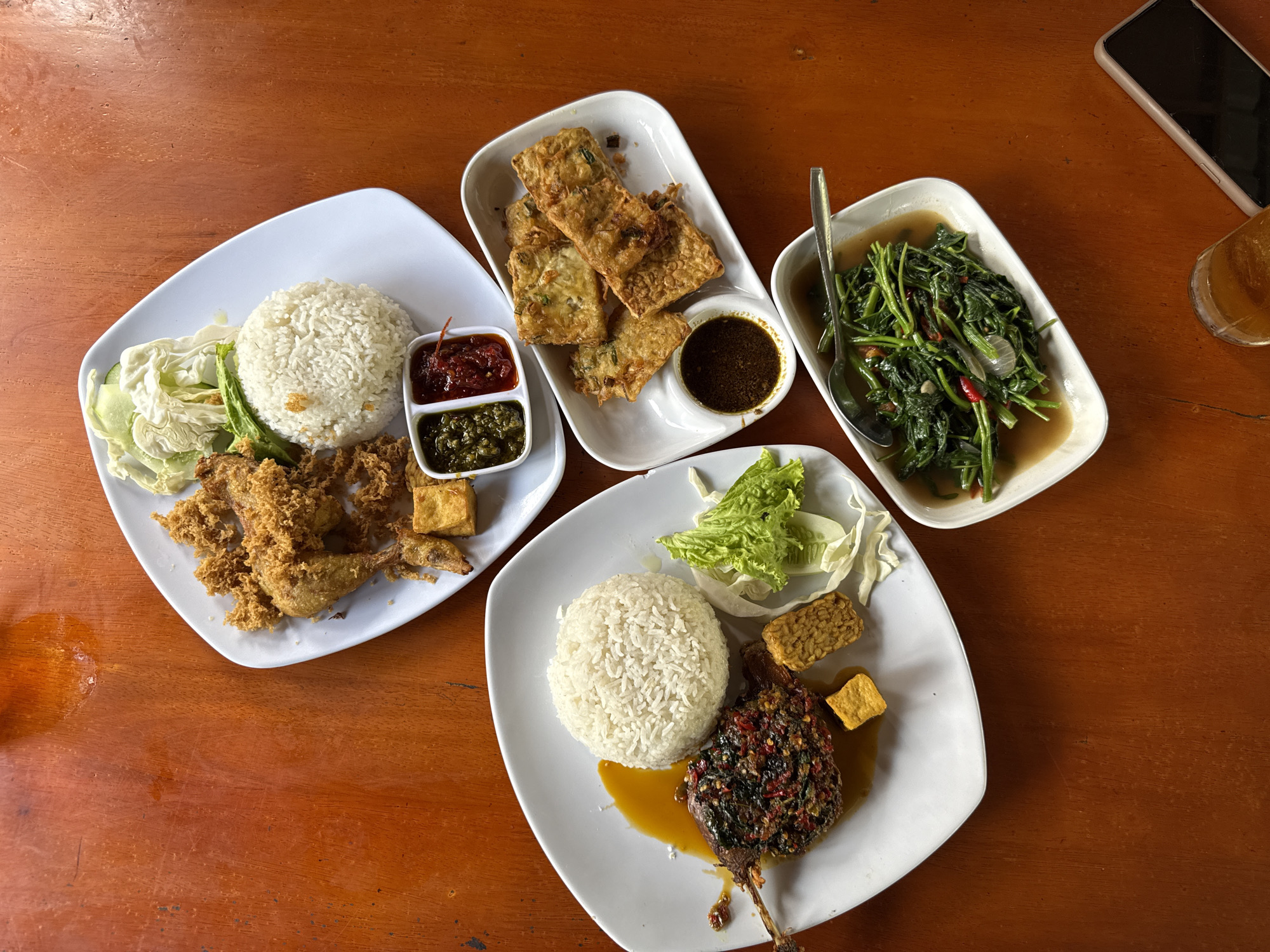 The image captures a spread of Indonesian dishes on a wooden table. Four white plates are arranged around the table. One plate contains fried chicken with rice, salad and two sauces. Another plate contains fried tofu with a dark sauce. A third plate holds water spinach with a spoon inside. The fourth plate has rice with duck and salad and tofu. A smartphone and a glass can be seen on the table. The scene is in Lhokseumawe, Indonesia.
