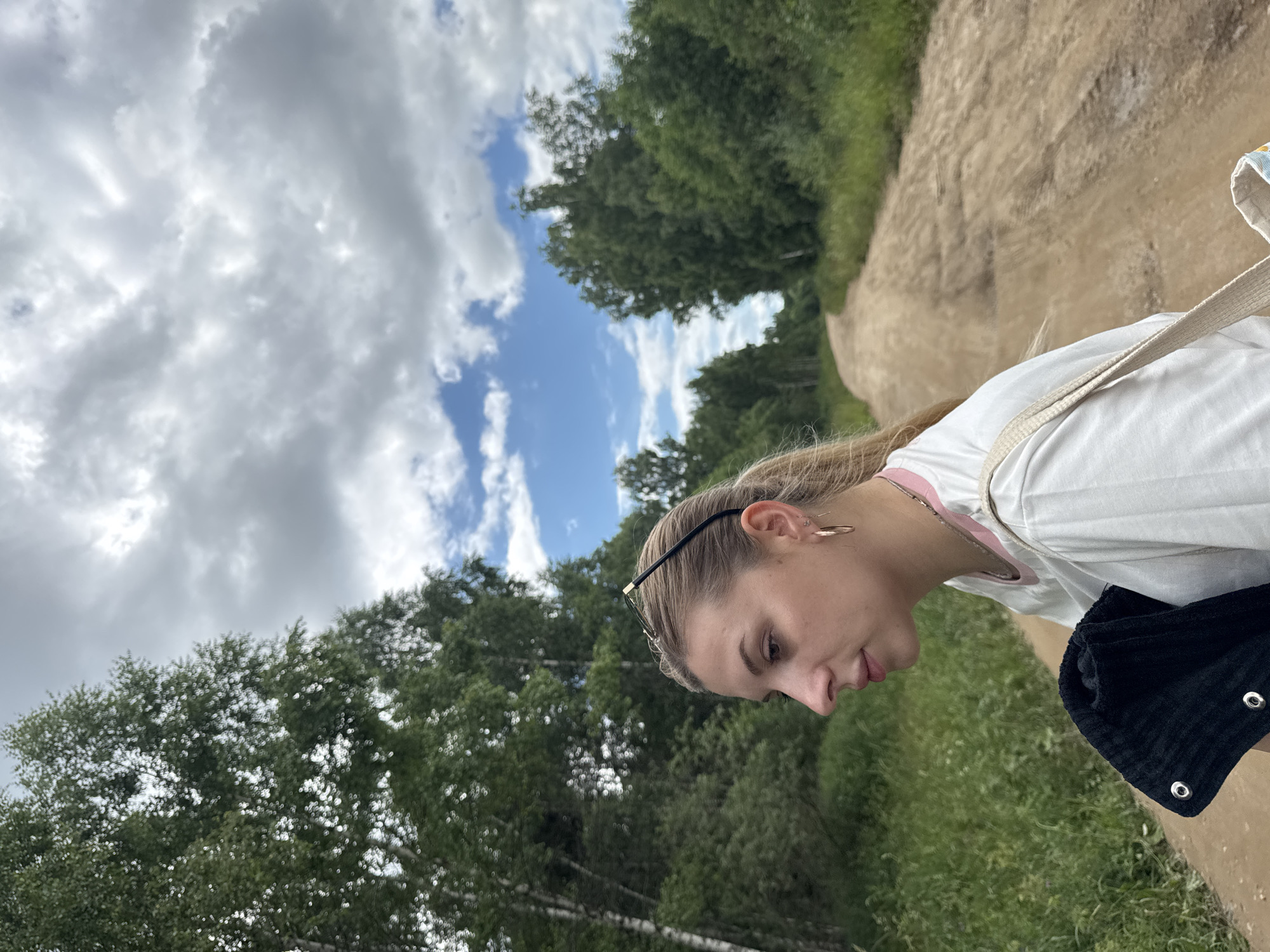 The image is a portrait of a young woman with blonde hair pulled back, wearing glasses and hoop earrings. She is wearing a white t-shirt with a pink collar. She is looking down and to the right, with a slight smile. The setting is outdoors, with a dirt path to the right and lush green trees and grass to the left. The sky is visible with scattered white clouds against a blue background, suggesting it is daytime with partly cloudy weather. A black bag is slung over her shoulder. The orientation of the image is rotated 90 degrees counter-clockwise. Based on the provided location context, the scene is likely in Krasnoe, Russia.