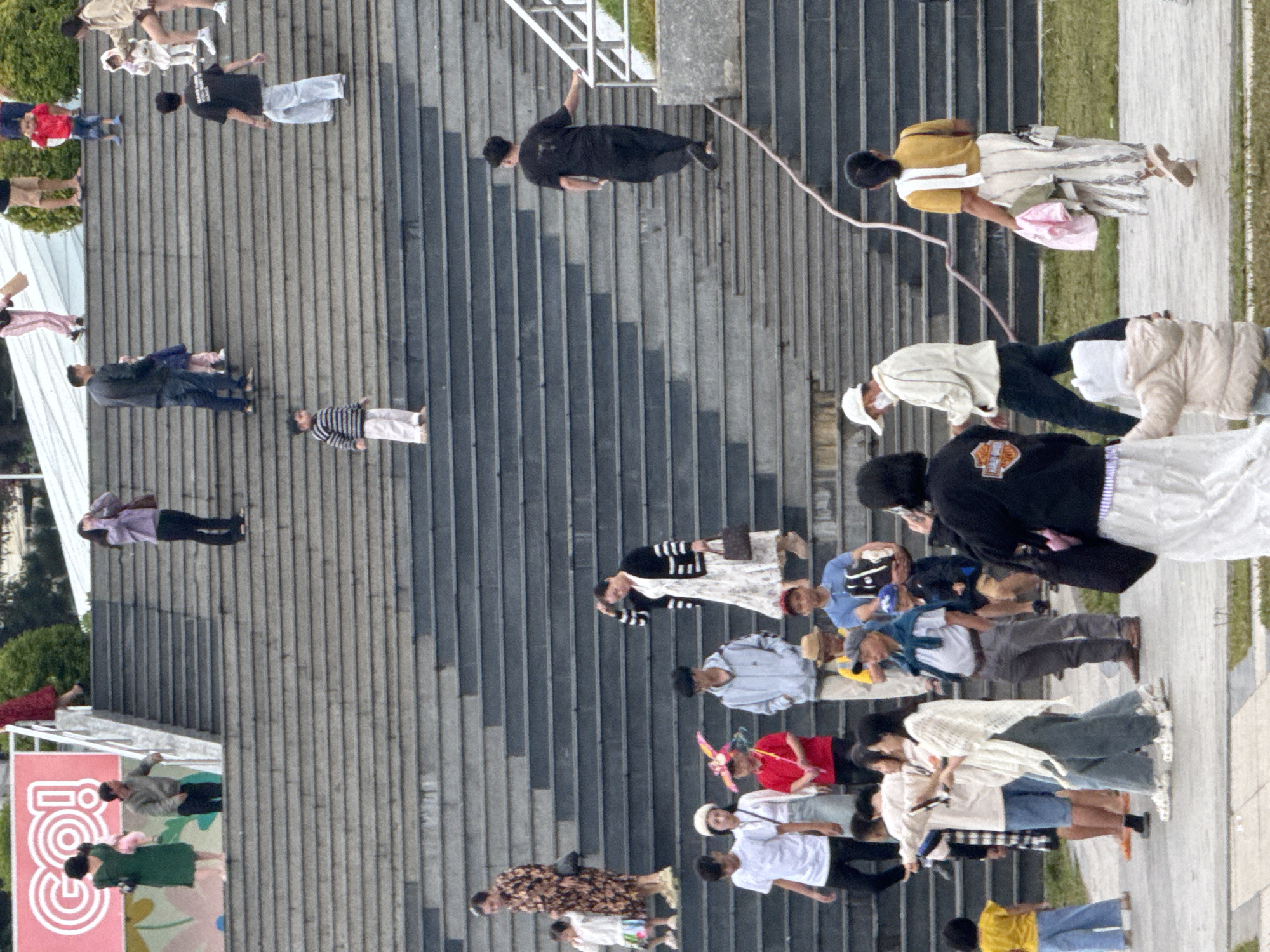 The image captures a wide outdoor staircase, likely part of a public space or monument in Da Lat, Vietnam, judging by the architectural style and general ambiance. The scene is populated with a diverse group of people of various ages, engaged in activities typical of a leisure outing.
In the foreground, several individuals are seen walking on a paved area at the base of the stairs. A child in a yellow shirt and blue pants is facing away from the camera. To their left, two women are walking together, one in a white, lacy shawl and the other in a black jacket. The woman in the shawl is holding a cell phone, seemingly taking a picture or recording a video. Another woman in a white skirt and a yellow top is walking away from the camera, carrying a pink garment and a purse.
Moving up the stairs, several groups and individuals are scattered across the steps. On a mid-level landing, a woman in a patterned dress walks with a child. Further up, a woman in a black and white striped sweater and a white dress is holding a handbag. A man in a black t-shirt and light-colored pants walks up the stairs, while another man in a dark jacket and jeans stands on a higher level.
Higher up, a man in a red shirt and shorts stands with a child in blue shorts. A family is visible near the top of the visible staircase, with a man in a dark shirt and jeans, a woman in a white top and shorts, and a child in a white outfit. On the upper levels, more individuals are scattered, including a person in a striped long-sleeved shirt and light pants, and another in a dark jacket and light pants.
The staircase itself is constructed with dark grey stone or concrete treads and risers. The steps are wide and appear to lead upwards towards a building or an elevated area not fully captured in the frame. To the left of the stairs, a large pink banner with white graphics, possibly an advertisement or signage, is partially visible. The background shows greenery and hints of a building with a white roof structure, consistent with Da Lat's landscape. The overall lighting suggests it is daytime, possibly overcast or with diffused sunlight.