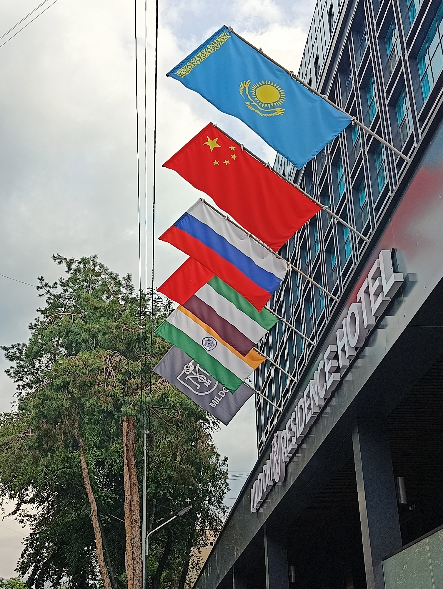 Several national flags are displayed vertically along the side of a modern building. The flags, from top to bottom, appear to be those of Kazakhstan, China, Russia, India, and another flag with a crest. The building has a facade of dark windows and a prominent sign reading "RESIDENCE HOTEL" in large white letters. A tall green tree stands to the left of the flags.