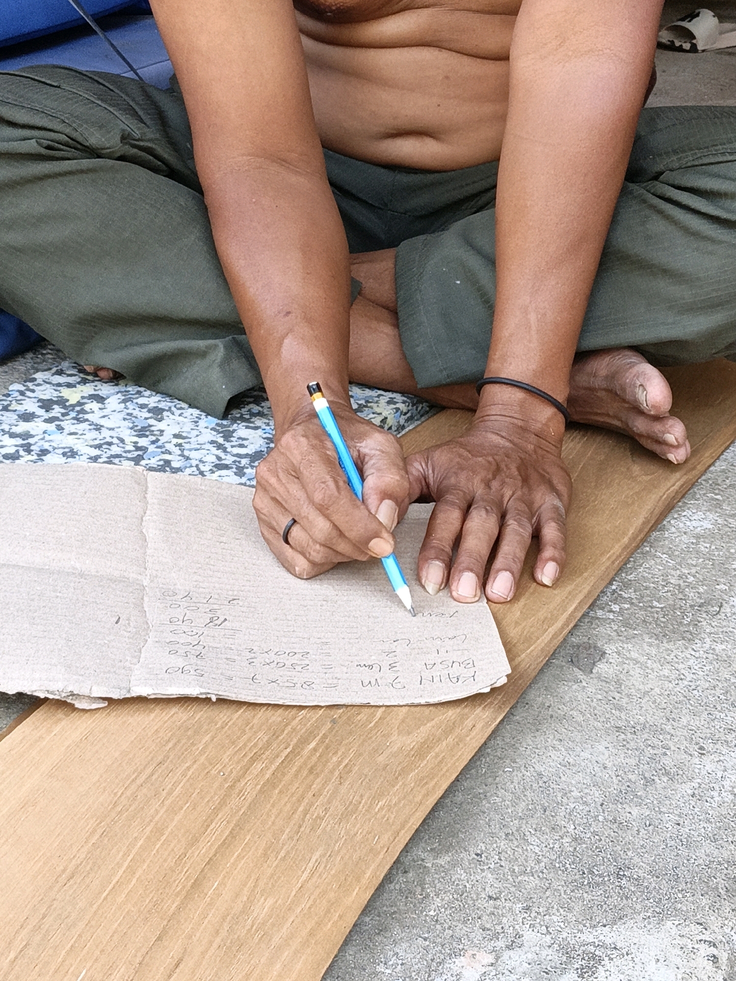 The image shows a person sitting on the ground in an outdoor setting. The person, whose torso is bare, is wearing green pants and has their legs crossed. They are holding a blue pencil in their right hand and are writing on a piece of cardboard. The cardboard has some calculations and handwritten text on it. To the left of the person, there is a blue object, possibly furniture, and a patterned surface. The ground appears to be concrete and wood. The scene is likely in Cikal, Indonesia, given the provided context. The person's hands show visible veins and skin texture, and a dark ring is visible on the ring finger of their left hand. Their feet are also visible, with one foot resting on the ground.