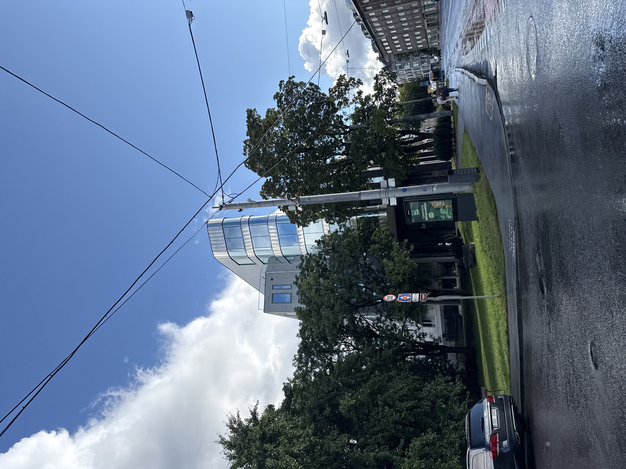 This is a street-level, medium-distance, eye-level shot taken in Tallinn, Estonia, on a day with scattered clouds and bright sunlight. The scene depicts a modern, curved glass-and-metal building on the left, contrasting with older brick buildings on the right. A wide, wet asphalt road with some puddles suggests recent rain. Overhead, power lines are visible against the blue sky. Trees line the sidewalk, and a parked dark-colored sedan is on the left side of the road. A few pedestrians are walking on the sidewalk towards the right. Traffic signs are present, including a "30" speed limit sign and a sign with a red circle and diagonal bar over a symbol depicting a pedestrian and a bicycle, indicating "no pedestrians and bicycles." A bus is visible further down the road on the right. The overall atmosphere is urban and functional, with evidence of recent precipitation.