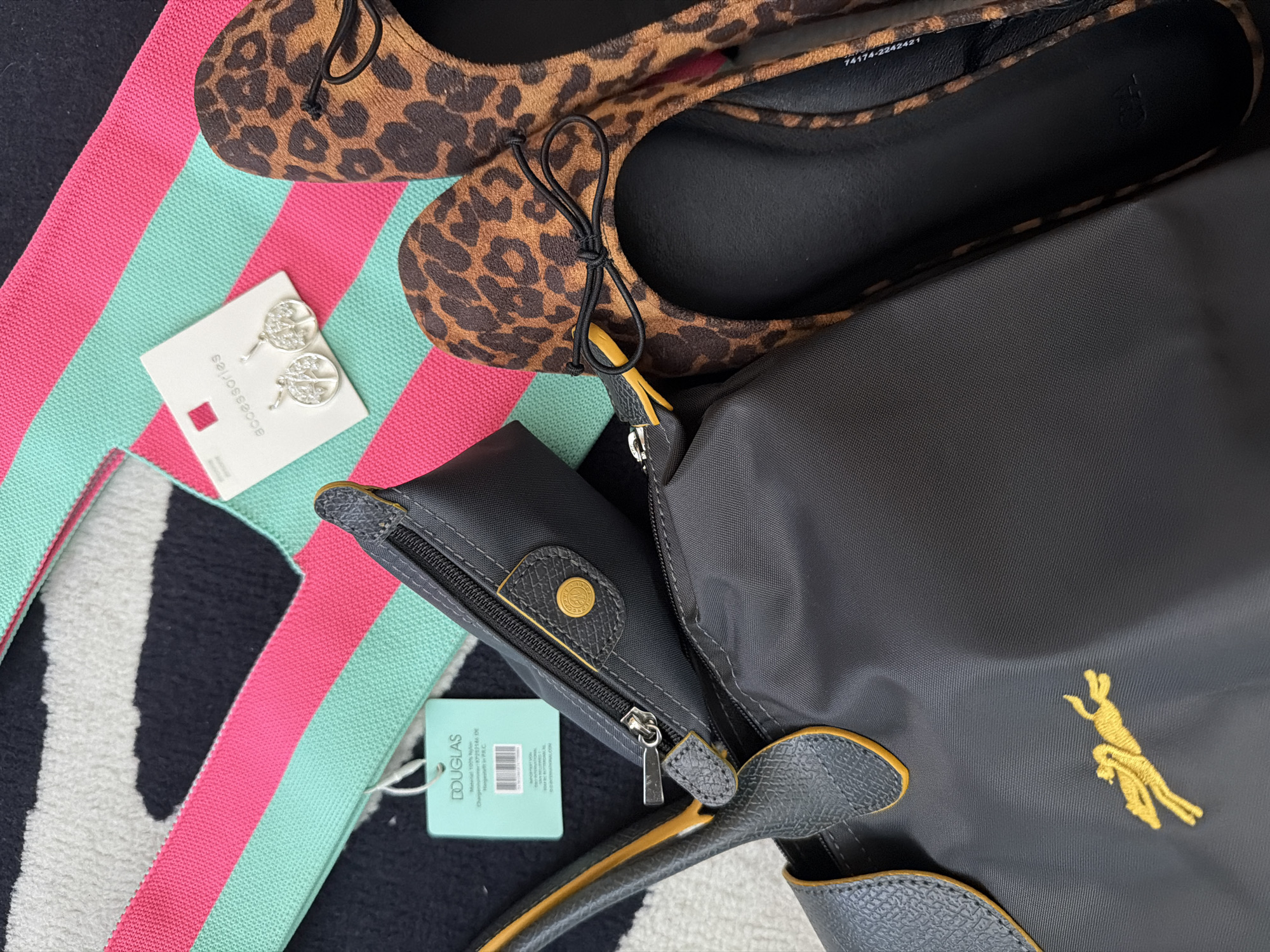 An overhead view displays a collection of personal accessories arranged on a multi-patterned surface. The background consists of a rug or textile featuring broad horizontal stripes of teal/mint green and hot pink, superimposed over a black and white abstract pattern.
In the foreground, a dark gray nylon tote bag, possibly a Longchamp Le Pliage, is partially open. It features a gold embroidered silhouette of a running horse or pony on its side, and dark gray textured faux leather trim with yellow piping along the edges of its handles. A small, dark gray zipped pouch or insert is positioned within the main compartment of the tote, revealing a silver zipper pull and a gold-colored button with an embossed design. A light blue rectangular tag from "DOUGLAS" is visible near the pouch, showing text including "Material: 100% Nylon" and "Hergestellt in P.R.C." (Made in P.R.C.).
To the right of the bag, a pair of women's ballet flats with a brown and black leopard print pattern are present. Each shoe features a small black bow on the vamp, and their inner soles are black. A partial white label with "1417A-2202421" is visible on one inner sole.
Resting on the striped background, positioned between the bag and the shoes, is a white cardboard display card labeled "accessories". Secured to this card are a pair of silver-toned dangle earrings, each depicting a "Tree of Life" motif. No individuals are present in the scene, and no actions or interactions are depicted beyond the static arrangement of objects. The environment is an indoor setting, likely a floor or flat surface where items have been laid out.