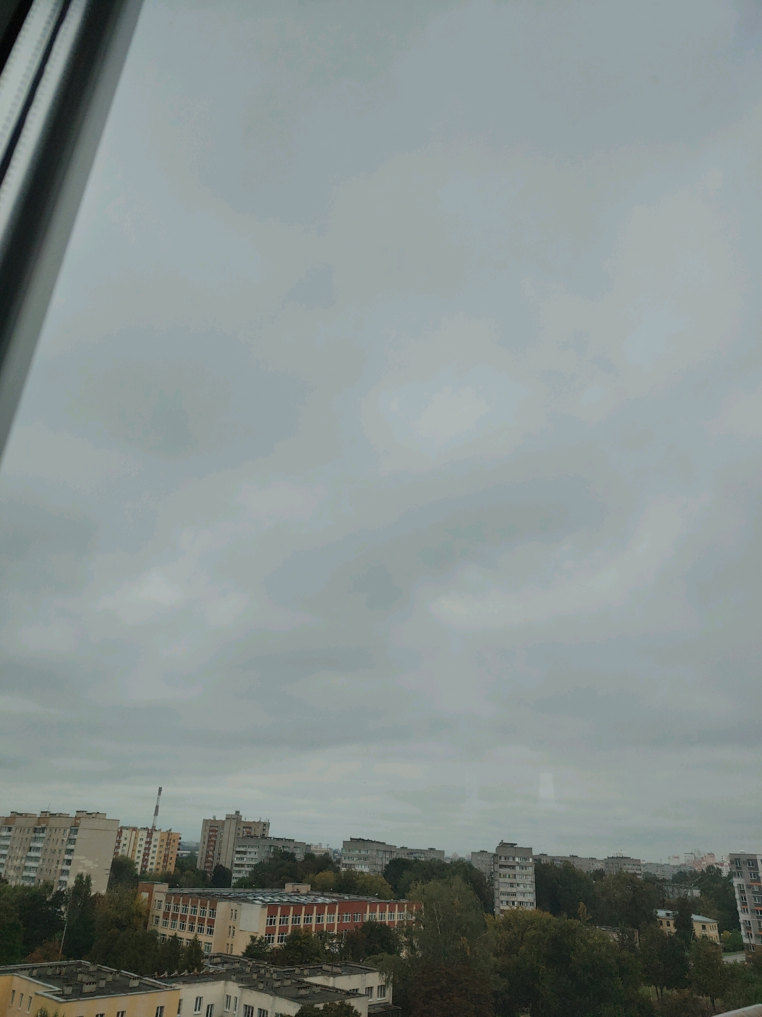 The image shows a city skyline under a cloudy sky, likely taken from an elevated position, such as a window. The perspective is from the left side of the frame, looking out towards the right. A window frame is visible on the left edge of the image.
In the foreground, on the left, are several buildings with flat roofs, suggesting residential or commercial structures. Moving towards the right and further into the distance, a variety of apartment buildings are visible. These buildings are of different heights and architectural styles, with some appearing to be older, multi-story concrete structures and others more modern. One building in the middle distance has a distinctive tall, red and white chimney or tower.
Green trees are interspersed throughout the cityscape, indicating the presence of parks or tree-lined streets. The overall impression is of a typical urban environment with a mix of residential buildings, infrastructure, and vegetation. The cloudy sky suggests overcast weather.
Given the context of Homyel, Belarus, this view likely depicts a residential area of the city, possibly a microdistrict, characterized by Soviet-era apartment blocks and surrounding green spaces.