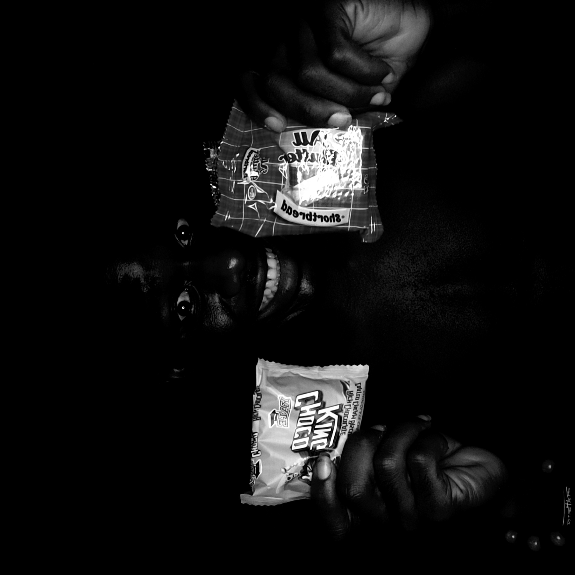 A black and white photograph depicts a person, likely lying down, smiling with teeth visible. Two hands hold packets of sweets above and below the person's face. The upper packet is checkered with indecipherable text, while the lower packet contains text reading "King Choco." The background is completely black. The location, Narayi, Nigeria, is mentioned but is not visually represented in the image.