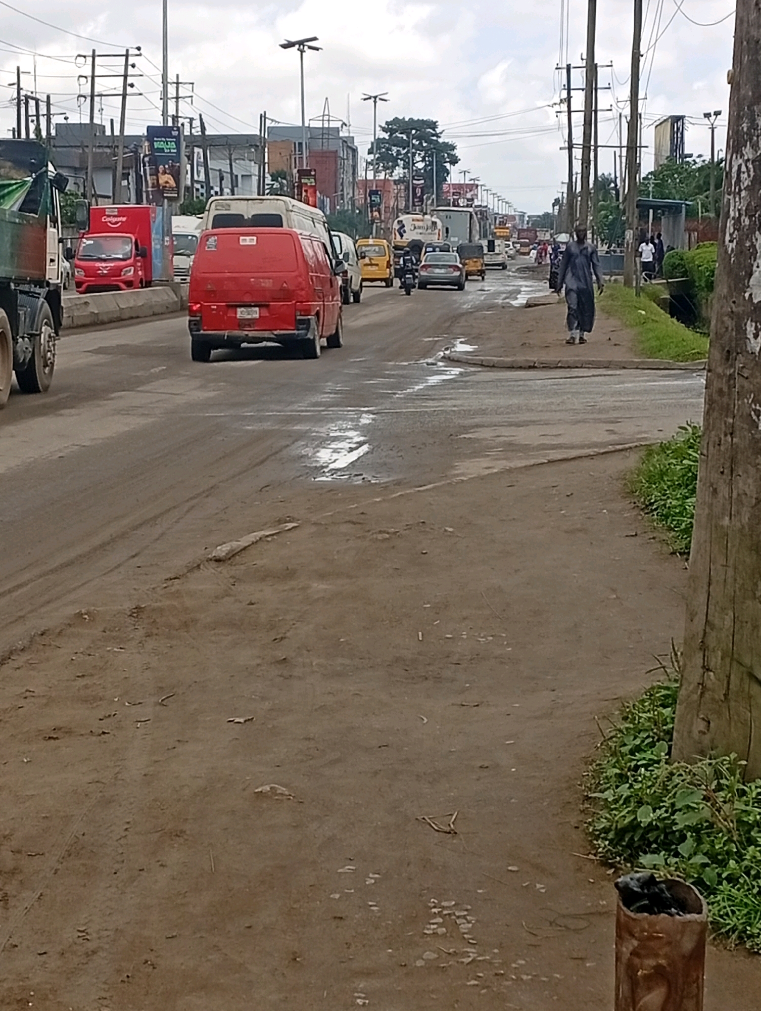 A medium-shot, eye-level, vertical video of a busy street scene in Ikeja, Nigeria shows a dirt road with multiple vehicles and a pedestrian. The foreground is dominated by the dusty, uneven ground and a wooden post on the right. In the mid-ground, several vehicles are on the road, including a red van with "Colgate" branding, a red car, and a red bus. A person in a dark blue robe walks on the sidewalk to the right of the road, facing away from the camera. Further down the road, more vehicles are visible, suggesting moderate traffic. The background features several multi-story buildings, billboards with advertisements, and utility poles with a network of wires, indicating an urban environment. The sky is overcast, and the overall lighting suggests it is daytime. The scene depicts a typical day on a bustling street in Ikeja.