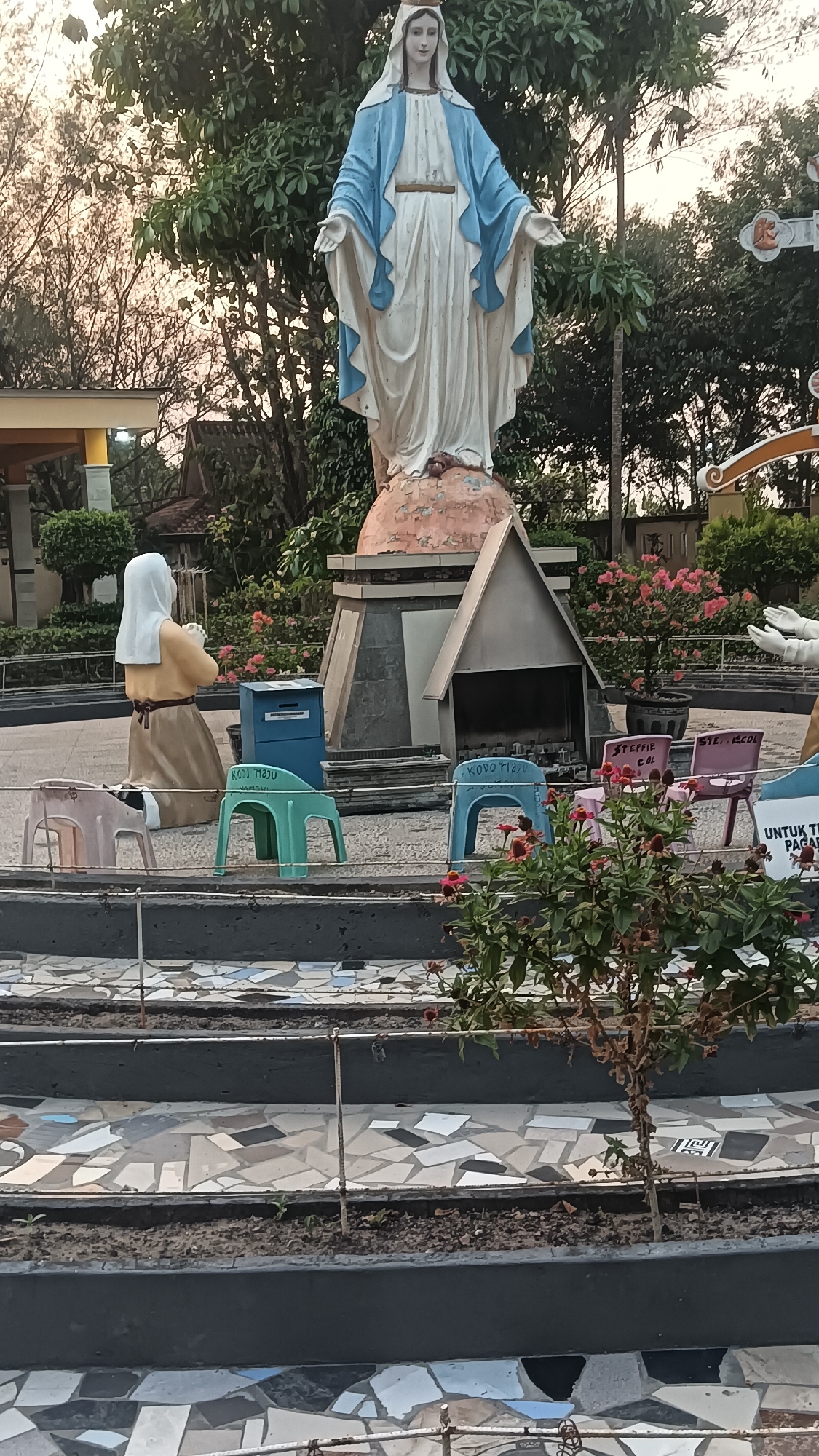A full shot captures a statue of the Virgin Mary in Dukuh, Indonesia. The statue, dressed in white and blue, stands with outstretched arms atop a pedestal. To the left, a kneeling statue of a woman in prayer faces away from the viewer. Several colorful plastic chairs are arranged in front of a small shrine with a slanted roof. A blue mailbox is positioned behind the kneeling statue. The area is decorated with flowering plants and trees, and the ground is paved with mosaic tiles. The scene appears to be an outdoor religious site.