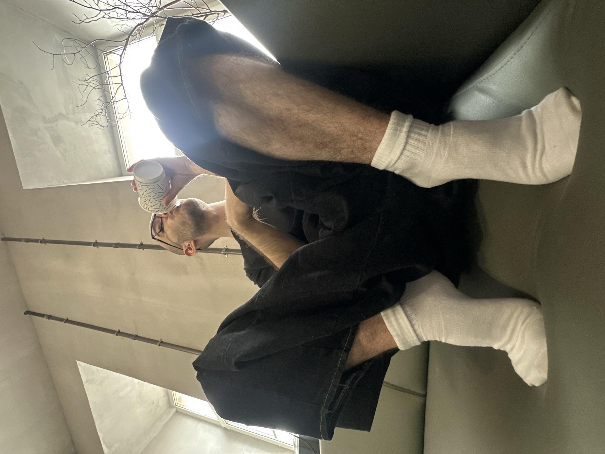 This image, captured from a low angle and rotated 90 degrees clockwise, depicts an adult male in an indoor setting, possibly in Moscow, Russia. The man has a light beard/stubble and wears glasses. He is seen with his head tilted back, drinking from a white cup adorned with black abstract patterns (lines and dots).
He is dressed in a dark, likely black, hooded top and loose-fitting black pants, paired with white crew socks. His legs are bent, and his feet are resting against a light grey surface, appearing to be a sofa or cushion.
The background reveals a modern interior with light-colored, possibly concrete or plaster, walls and ceiling. Bright natural light streams in from an unseen window in the upper left, creating a strong highlight and lens flare. Bare tree branches are visible against this bright light, suggesting either an outdoor view or a decorative element. Two parallel metal pipes are mounted on the wall/ceiling in the mid-ground. The scene is well-lit, indicating it is daytime. The man's expression is neutral, focused on drinking. No visible text is present.