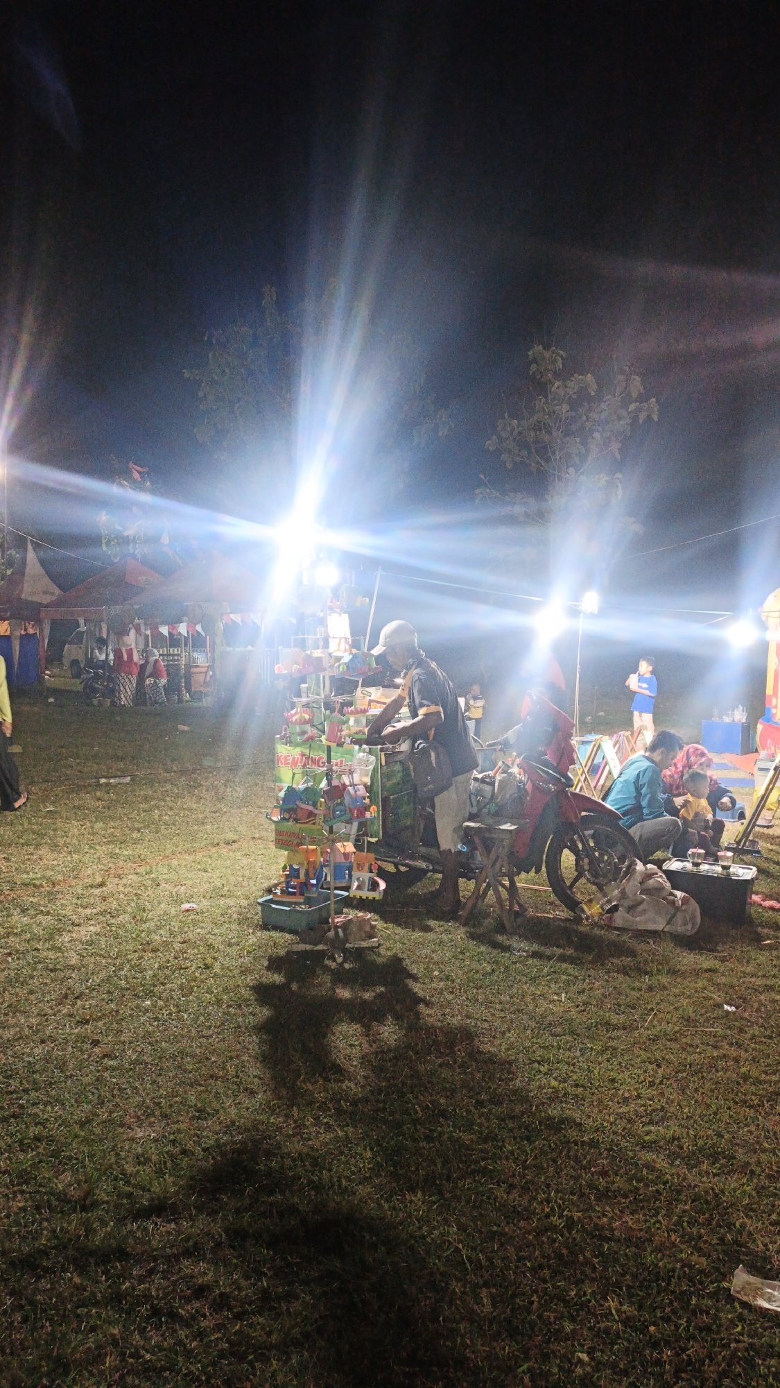 This is a night time, medium-shot, vertical, full-color photograph of a street vendor's stall in Ketemasdungus, Indonesia. The scene is illuminated by bright, harsh overhead lights creating lens flares. In the foreground, a man wearing a white cap, a dark blue shirt with yellow trim, and light-colored shorts, stands behind a mobile stall laden with colorful toys. The stall appears to be mounted on a cart. To the right of the stall is a red motorcycle with a kickstand down. A bundle of items wrapped in plastic is on the ground next to the motorcycle, along with a small wooden stool. A dark shadow of the vendor and his stall is cast on the grass in the foreground. In the middle ground, to the left of the vendor, are several tents with lights, suggesting a marketplace or fair. People can be seen near the tents. To the right of the motorcycle, two individuals are seated on the ground, one wearing a blue shirt and the other a headscarf. The ground is covered in green grass, and the overall impression is of a lively outdoor evening event.