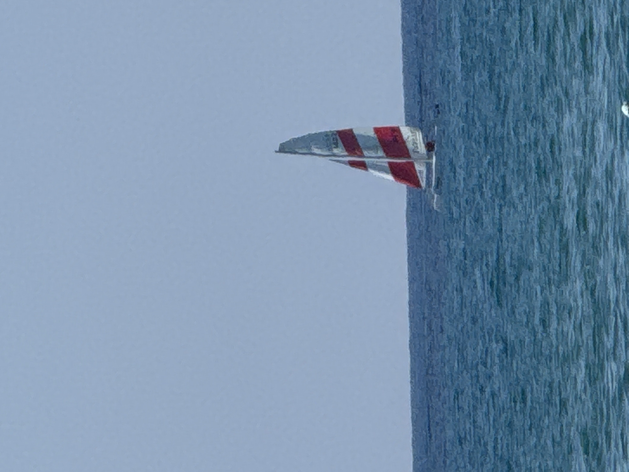 A lone sailboat with red and white striped sails navigates the blue waters off the coast of Manavgat, Turkey, under a clear, pale blue sky. The horizon line is sharply defined, separating the calm sea from the sky. The sailboat is positioned in the right half of the frame, with its sails fully extended, indicating it is actively sailing. The water exhibits subtle ripples, suggesting a gentle breeze. There are no people visible on the boat or in the surrounding water. The image captures a serene maritime scene with no discernible human activity beyond the single vessel.