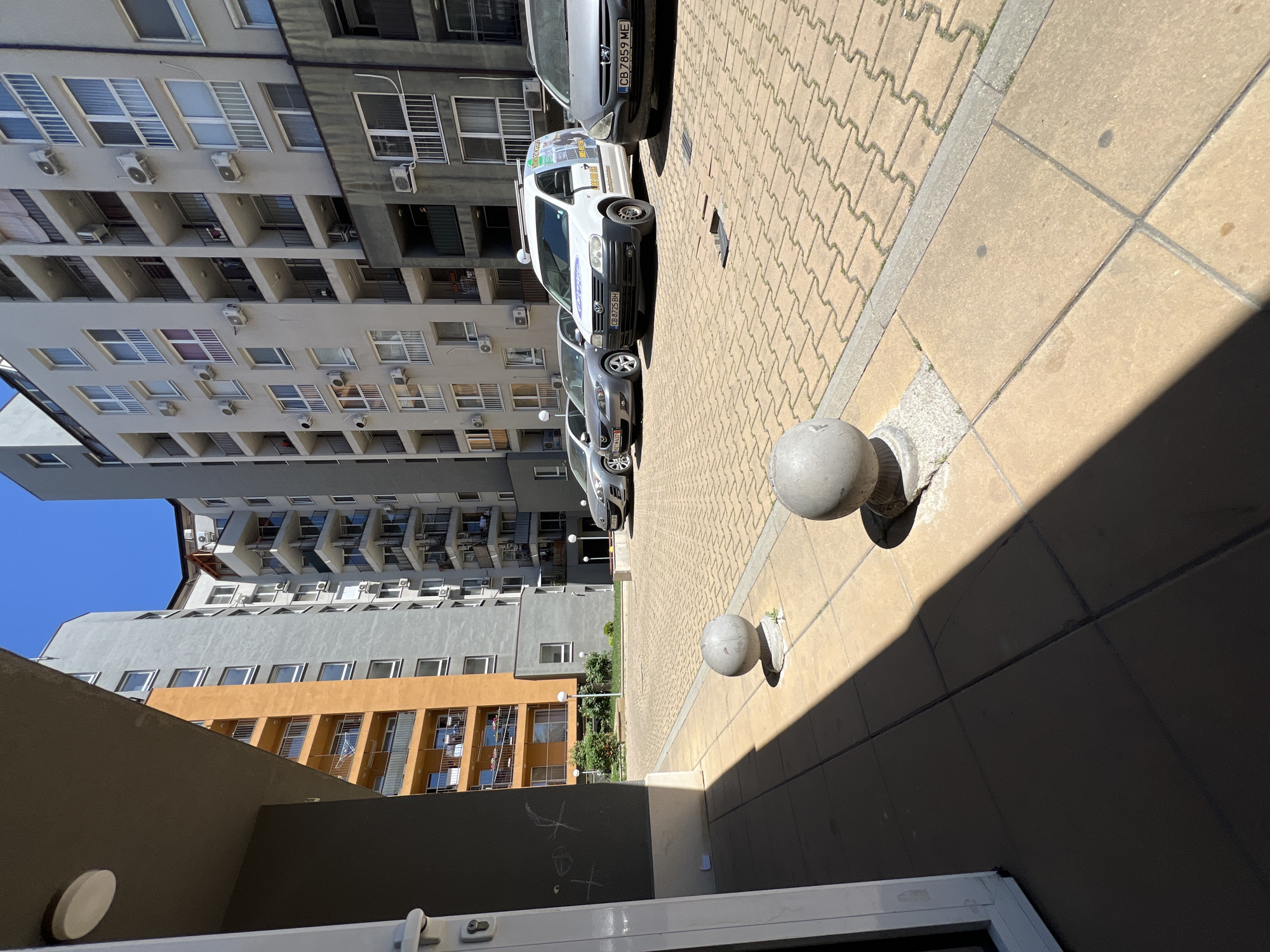 A street scene in Sofia, Bulgaria, presents a view upwards towards tall apartment buildings under a blue sky. A row of parked vehicles is visible along the street, including a white van. Concrete bollards line the edge of the sidewalk. The ground is paved with rectangular patterned paving stones, transitioning into larger square paving stones closer to the viewing point. The perspective is somewhat tilted. The foreground shows part of a building's entrance, with a white door frame.