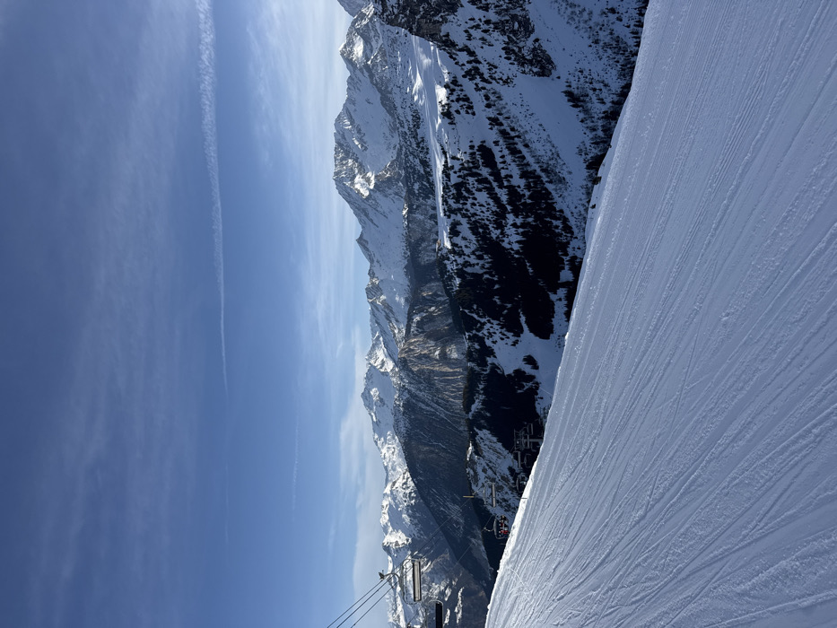 The image depicts a panoramic winter mountain scene in Courchevel, France. In the immediate foreground, a wide, groomed ski slope covered in fresh snow exhibits visible corduroy tracks, sloping downwards from the upper left towards the lower right. In the mid-ground, a section of a ski lift cable and two empty chairs are visible ascending towards the upper left, and further down the slope, another ski lift tower with a single chair is present. The landscape beyond the slope consists of a deep valley with steep, snow-covered terrain, interspersed with dark green coniferous forests and prominent rocky cliffs. The background features a vast range of towering, snow-capped mountains illuminated by bright sunlight, with their rugged peaks defined against a clear, deep blue sky. A thin, white contrail stretches horizontally across the upper sky. No specific individuals are discernible on the slope or within the ski lift chairs.
