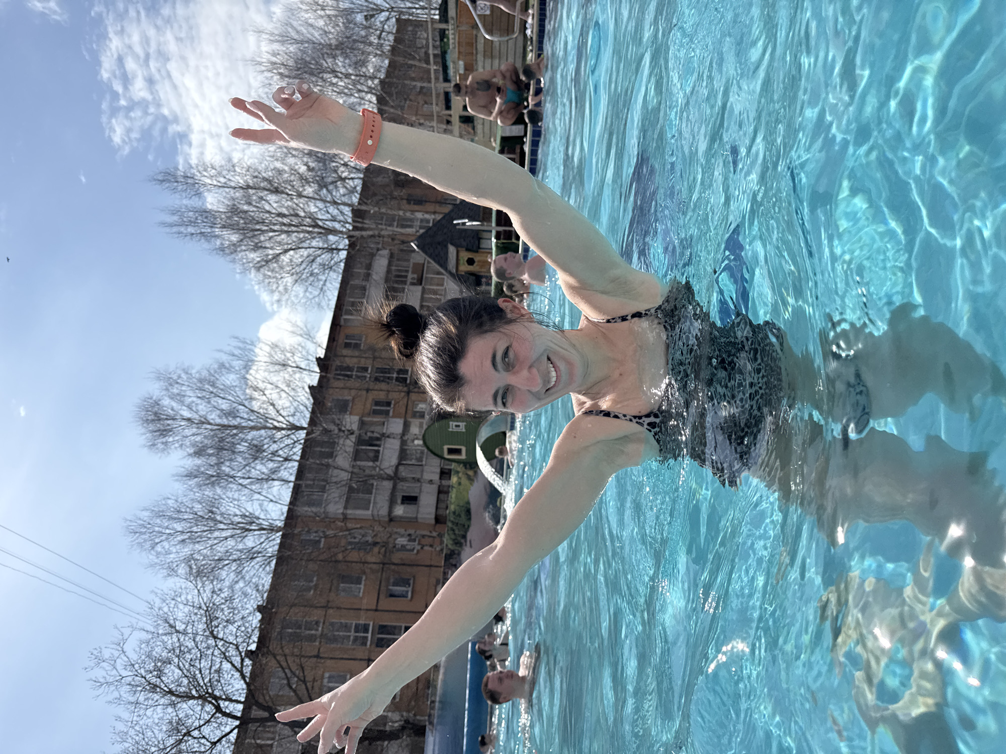 A woman is in a pool, arms outstretched, wearing a patterned swimsuit and a bracelet. The location is Votkinsk, Russia. A building and trees are visible in the background. Other people are in the pool. The water is clear.