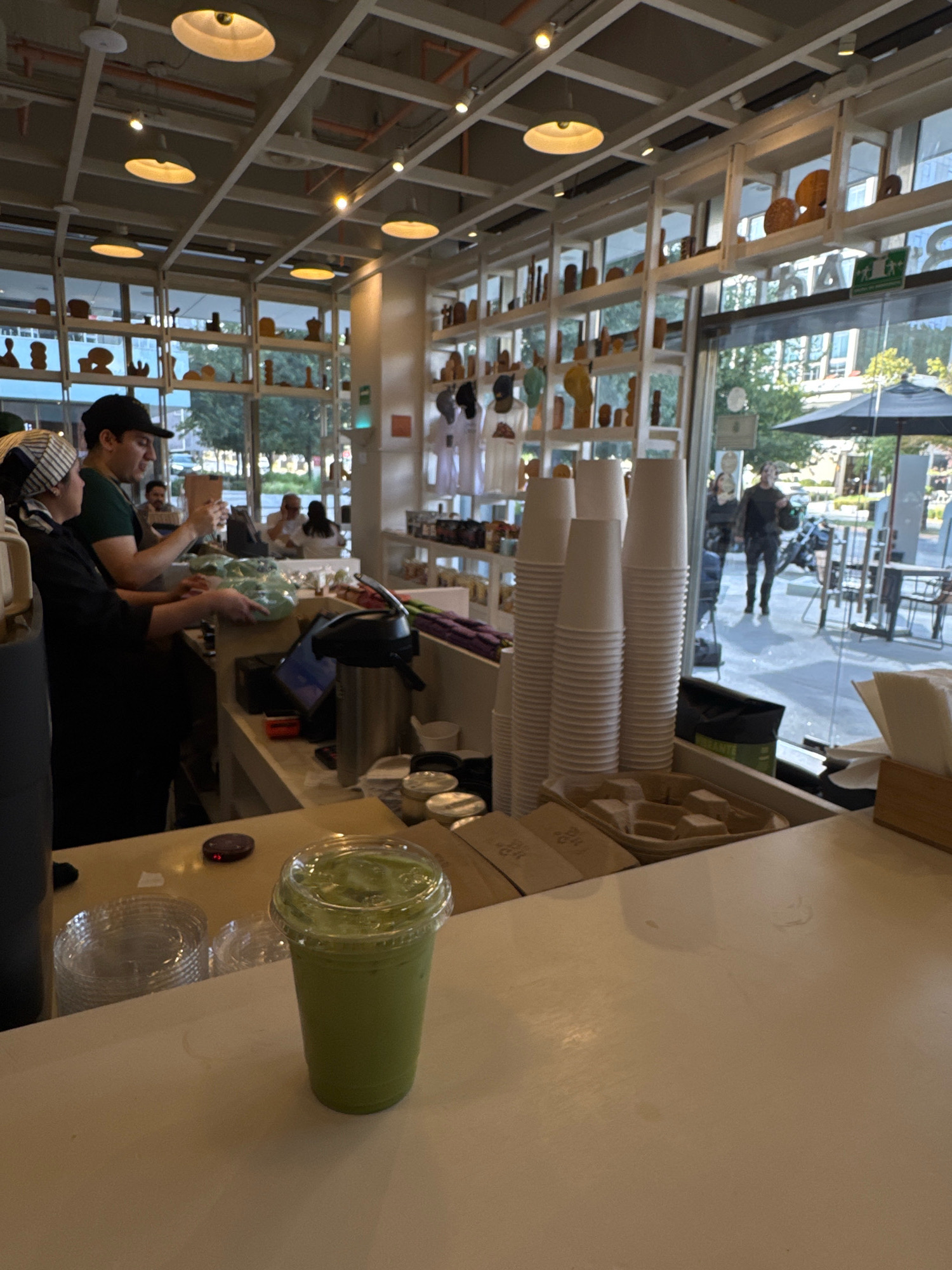 The image depicts the interior of a brightly lit cafe in San Pedro Garza García, Mexico. In the foreground, a clear plastic cup filled with a vibrant green beverage, likely a matcha latte, sits on a white countertop. To the left of the drink, stacks of clear plastic cups and a small red object are visible on the counter. Behind this, a man wearing a black cap and a green shirt is interacting with a woman dressed in a black apron and a striped headscarf, who is handling what appears to be a bundle of leafy greens. They are positioned behind a counter with a digital screen. In the background, through large windows and shelves displaying various decorative items and products, a street scene is visible. People can be seen walking outside, and outdoor seating with umbrellas is present. The cafe features a modern industrial ceiling with exposed beams and multiple pendant lights.