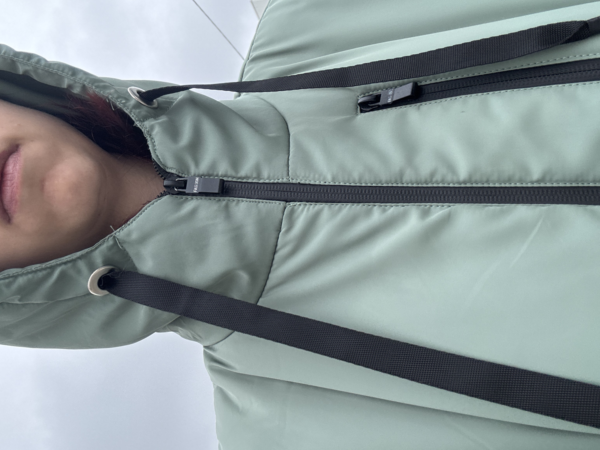 A close-up, chest-up shot shows a person wearing a light green hooded jacket. The jacket features a prominent black zipper down the front, with a smaller zippered pocket on the right side of the chest. Black drawstrings with metal grommets are visible on the hood. The word "FASHION" is printed in white on the zipper pull of the main zipper and on the smaller pocket's zipper pull. The background is a muted, overcast sky. The scene is set in an outdoor urban environment.