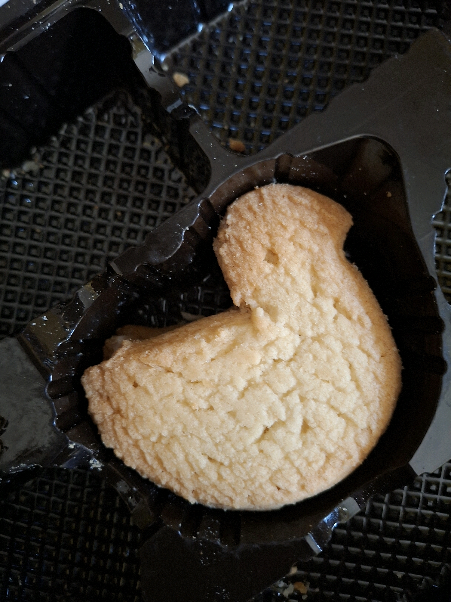 A close-up, top-down shot shows a single, light-brown, star-shaped cookie sitting in a black plastic tray. The cookie has a textured surface with visible cracks and indentations, suggesting it might be a shortbread or butter cookie. The plastic tray has a grid pattern on its surface and a somewhat glossy finish. There are a few small crumbs scattered around the cookie and on the tray. The lighting is somewhat harsh, creating shadows and highlights on the cookie and tray. The image appears to be taken indoors, possibly in a kitchen or dining area. There is no indication of the city of Lumajang, Indonesia.