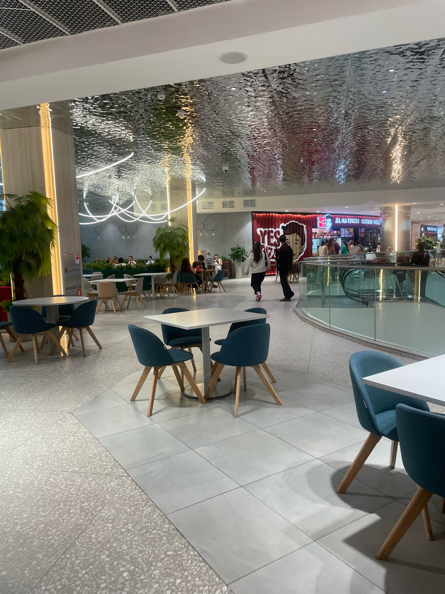 The image depicts an indoor seating area within a modern shopping mall, likely in Sochi, Russia, given the signage. The ceiling features a distinctive metallic, rippled surface that reflects the lighting. In the foreground, several tables with white tops and teal chairs are arranged on a light-colored tiled floor. To the left, a large potted plant with palm-like fronds stands near a pillar with embedded vertical lighting. In the background, a food court area is visible with more seating and a prominent "YES PIZZA" establishment. Several individuals are present, some seated at tables and others walking through the area. An escalator is also visible to the right of the food court, indicating a multi-level structure. The overall atmosphere is bright and commercial.