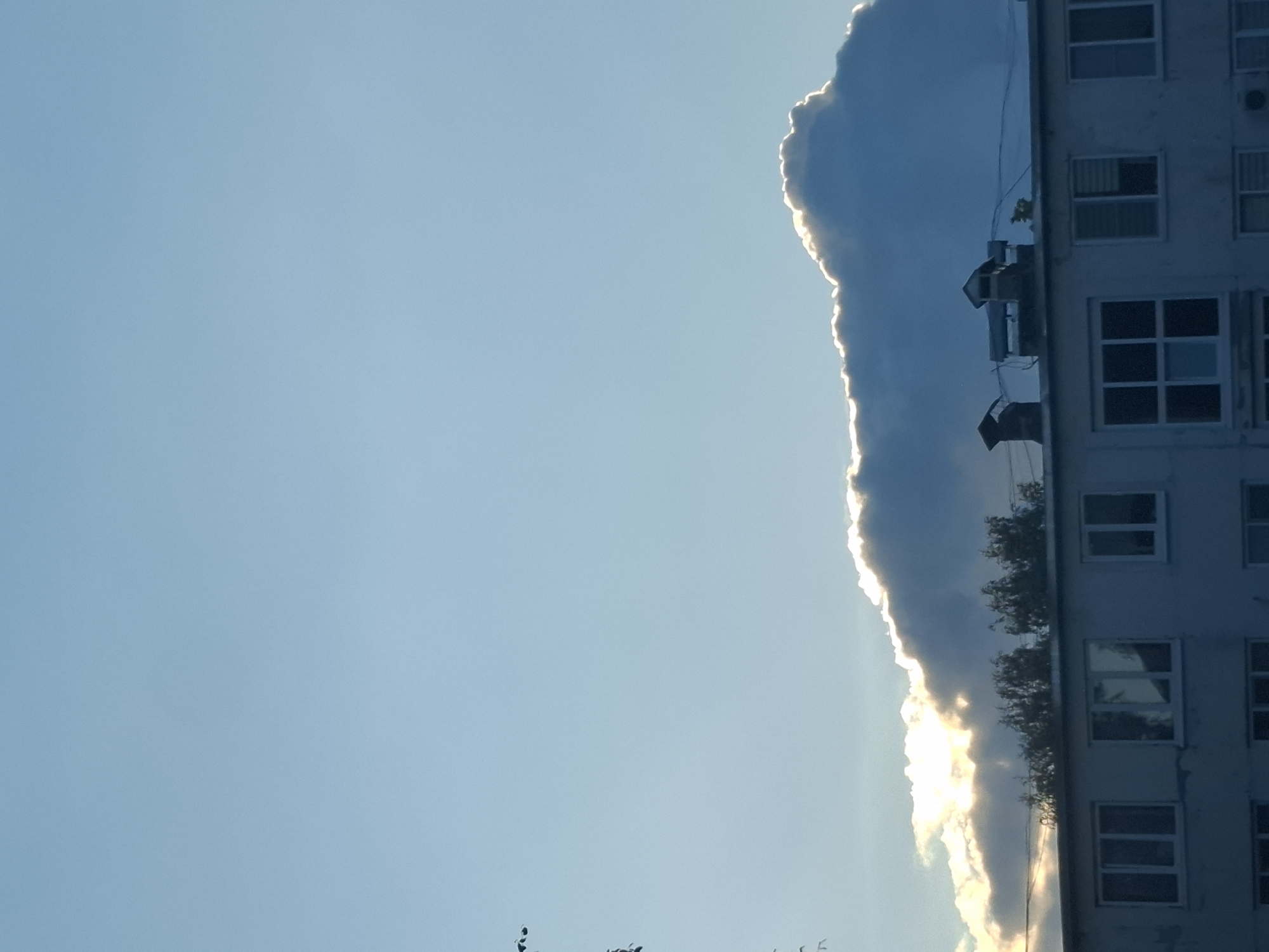 The image captures a portion of a multi-story building situated in Chaikovsky, Russia. The building's facade, with its rows of windows, is visible on the right side of the frame. The sky takes up the majority of the left side, filled with a dramatic cloud formation that is dramatically lit from behind, creating a bright, illuminated edge. The sun appears to be low in the sky, likely during sunrise or sunset, casting a warm glow on the cloud. The overall mood is serene and perhaps a bit awe-inspiring due to the striking cloud display. There are no people or specific activities depicted, but the scene suggests an ordinary urban setting with a beautiful natural phenomenon occurring. The time of day is indicated by the strong backlighting of the clouds.