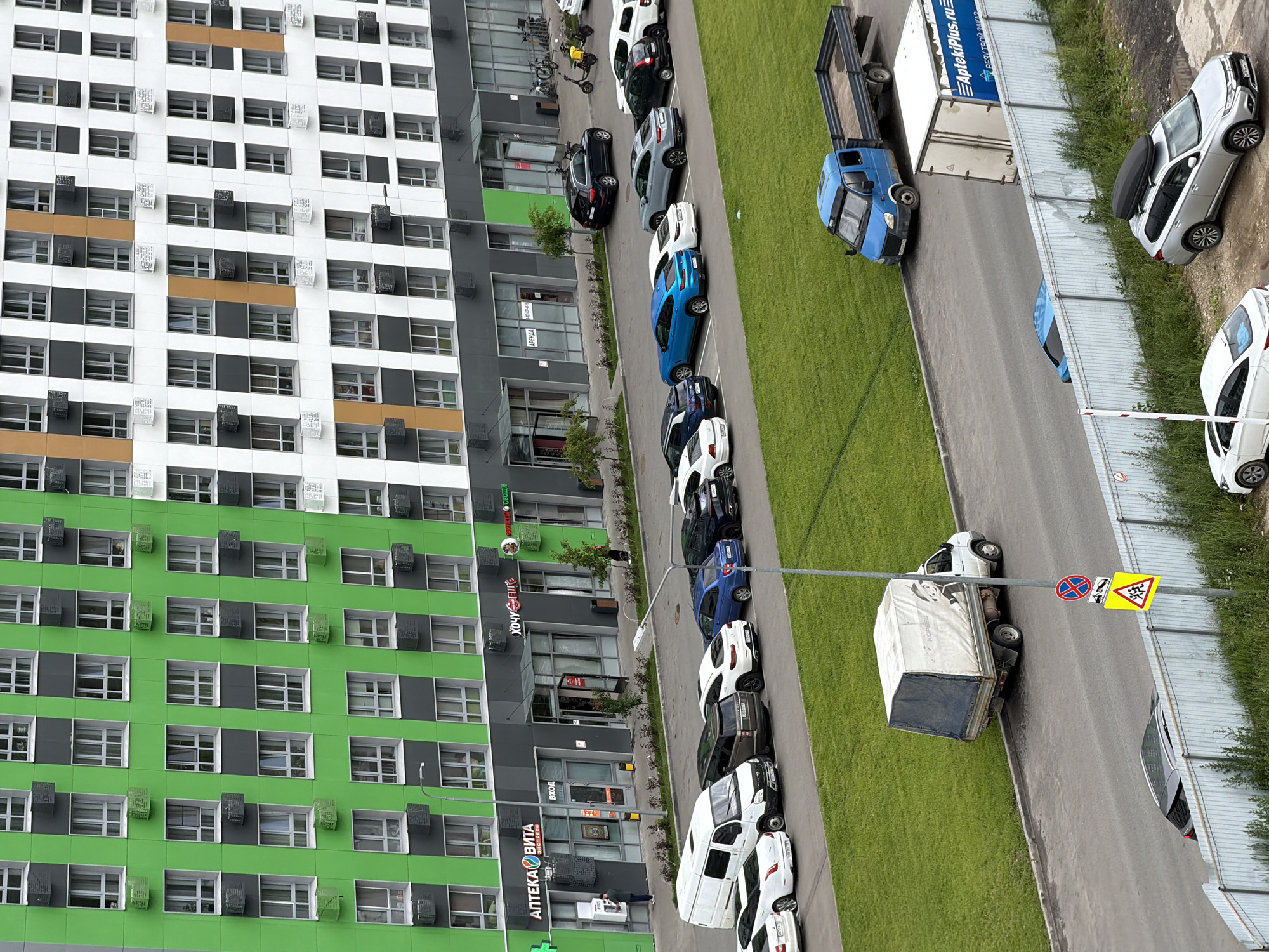 An exterior, eye-level, medium shot captures a row of apartment buildings with green and white facades, accented with orange and dark gray. Below the residential units, the ground floor features a commercial strip with several storefronts, including "APTEKA VITA EXPRESS" and "HOUCY BHIT." A person stands at an ATM outside the pharmacy. A paved area in front of the buildings is lined with parked cars, including a white van, a black SUV, a blue sedan, and several white and silver cars. Beyond the parked cars, a patch of green grass separates the road from the buildings. A white box truck with a blue tarp is parked on the side of the road, and a blue flatbed truck is further down the street. In the foreground, a white metal fence runs along the edge of the road, with a yellow triangular sign depicting children playing.