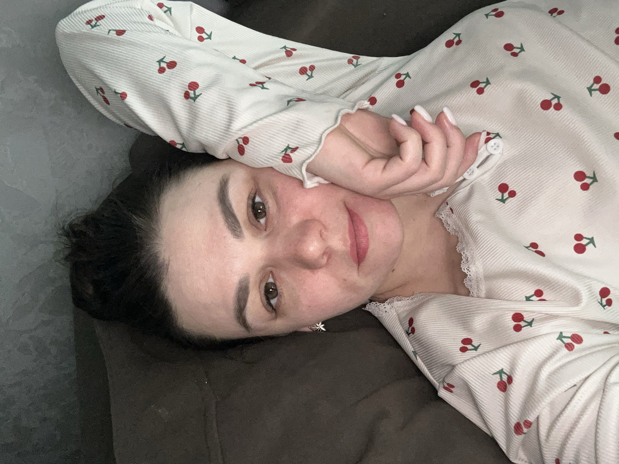 A woman is lying on a dark brown surface, possibly a couch or bed. She is wearing a white, long-sleeved shirt with a cherry pattern. Her dark hair is pulled back, and she is wearing an earring. Her hand is near her face. The image appears to be taken indoors. The location is not evident from the visual information and there is no way to determine if this image was taken in the city of Chaikovsky, Russia.