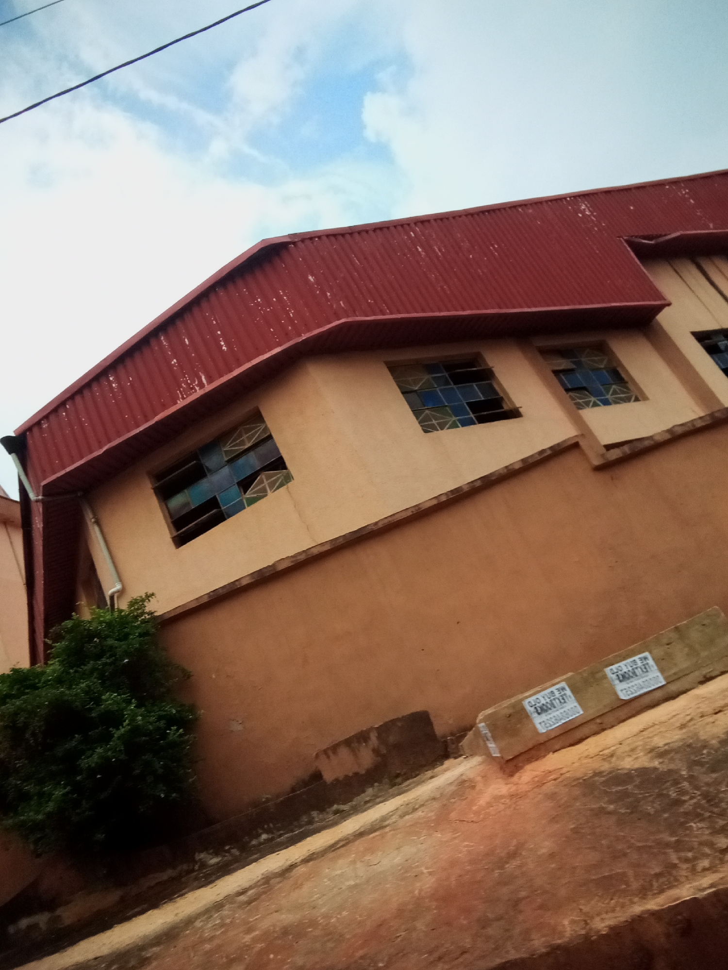 This is an eye-level, outdoor, medium shot image of a building with a red corrugated metal roof in Oko Town, Nigeria. The building has beige-colored walls with several windows featuring stained glass. There is a large green bush in the lower left corner of the image. A sloping, reddish-brown pathway leads away from the building. Two pieces of wood with labels are resting on the pathway in the lower right quadrant of the image. The sky above is partially cloudy, with some blue visible. A thin black wire stretches across the top of the frame.