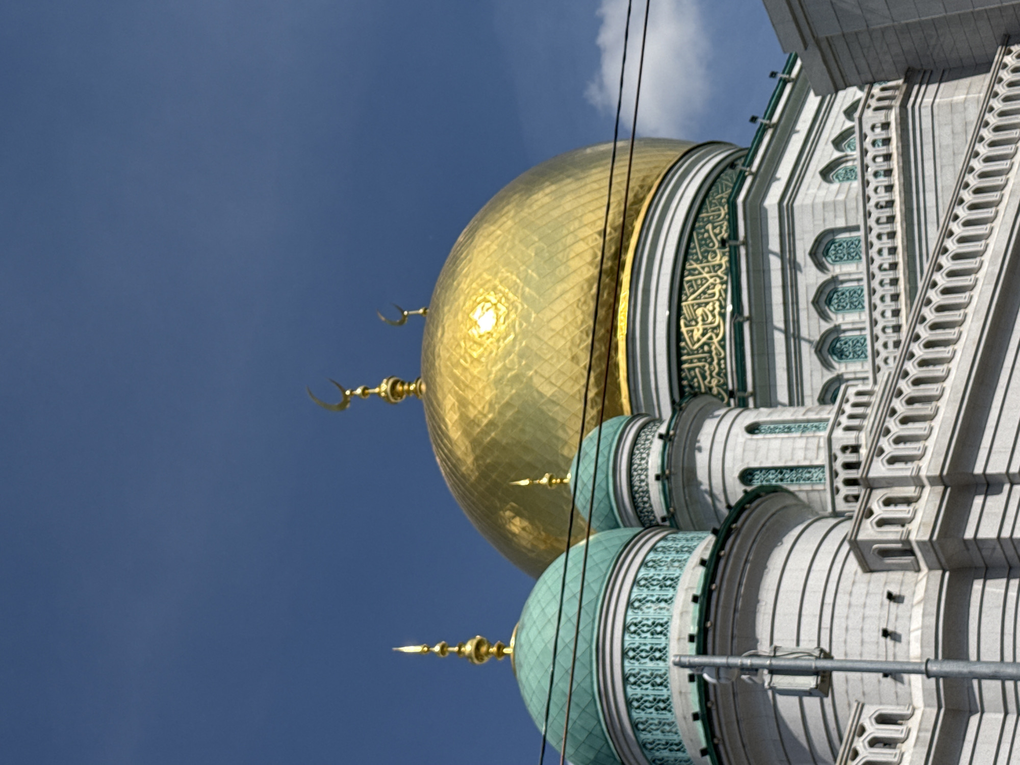 The image shows a section of the Moscow Cathedral Mosque in Moscow, Russia, viewed from a low angle. It features a large golden dome at the top, with a smaller green dome beneath it. Both domes have golden crescent moon symbols. The building's exterior is white with green and gold decorative elements, featuring intricate patterns and Arabic calligraphy. A clear blue sky serves as the background. Overhead power lines are visible against the sky.
