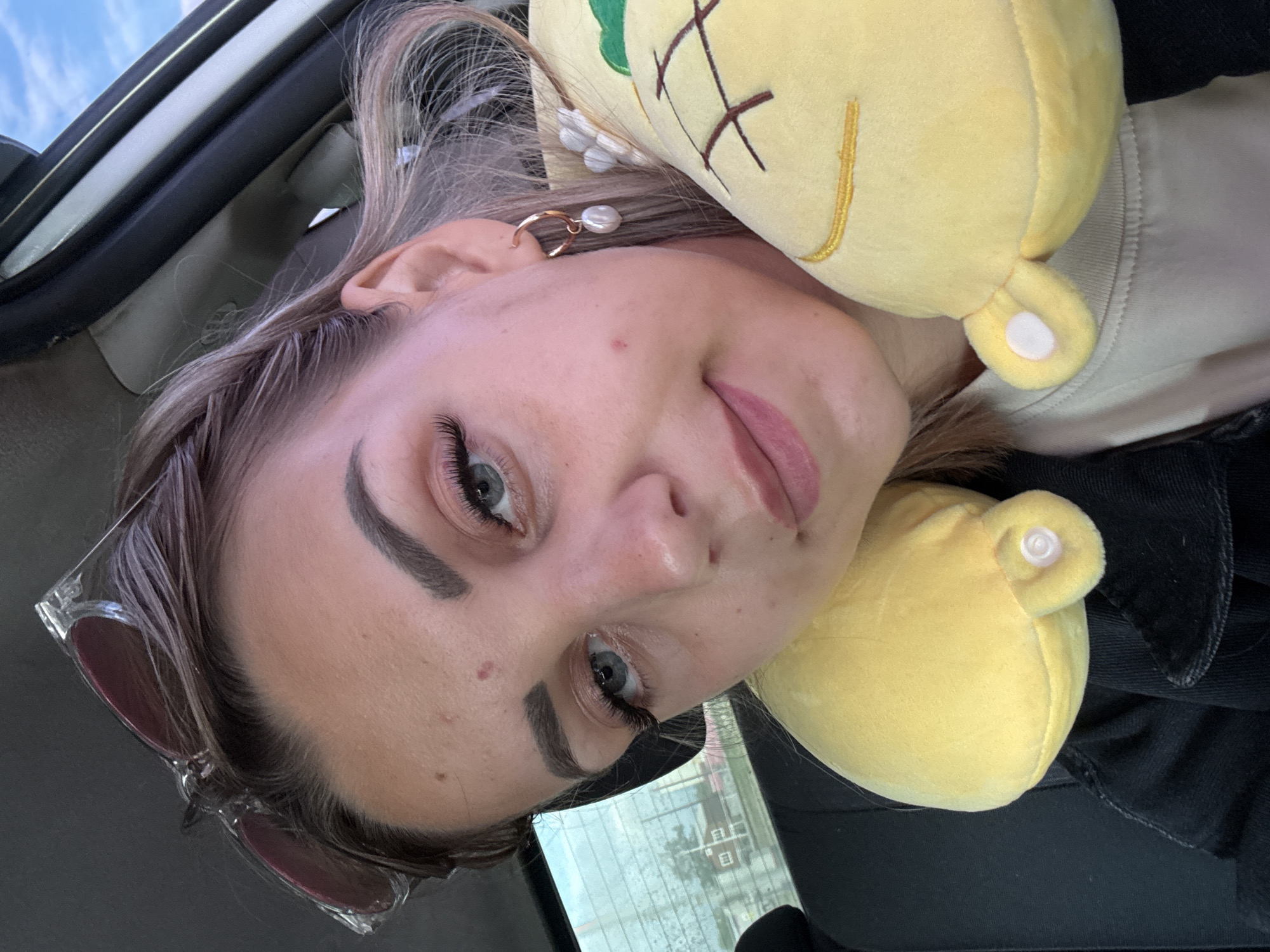 A young woman with blonde hair is in the passenger seat of a car, leaning on a yellow pineapple-shaped travel pillow. She is looking at the camera, her head tilted slightly. Sunglasses are perched on top of her head. She is wearing a black jacket over a light-colored shirt, and a pearl earring is visible on her right ear. The interior of the car is visible, as is a blurry outdoor scene through the window, suggesting the car is in motion or parked. The image appears to have been taken in Yuzhno-Sakhalinsk, Russia, based on the provided context.