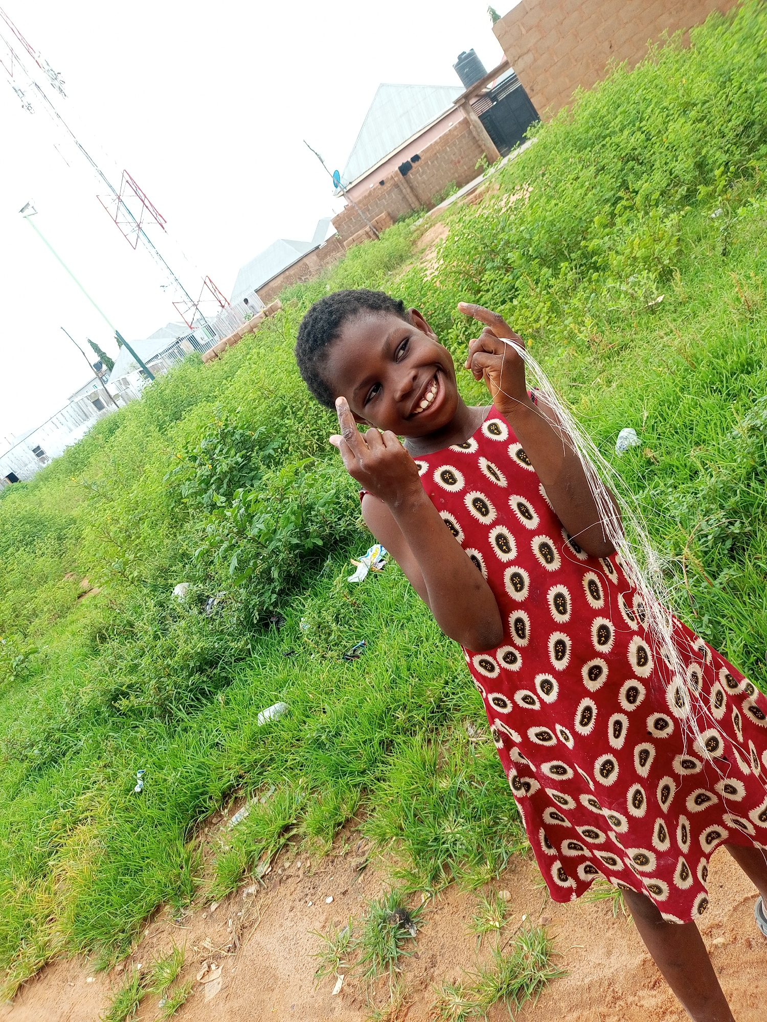 In Tempure, Nigeria, a young girl with dark skin and short black hair smiles at the camera. She wears a red dress with a white and black patterned design. Her left hand is raised, and she appears to be holding some white string. Her right hand is also raised, with her index finger extended upwards and her thumb extended horizontally, forming a gesture. The girl stands in an outdoor environment with lush green grass covering a slope. Behind her, a few buildings with light-colored roofs are visible, and in the background, a tall metal tower with red and white sections stands against a bright, overcast sky. The ground in the foreground is reddish-brown dirt with some sparse green vegetation.