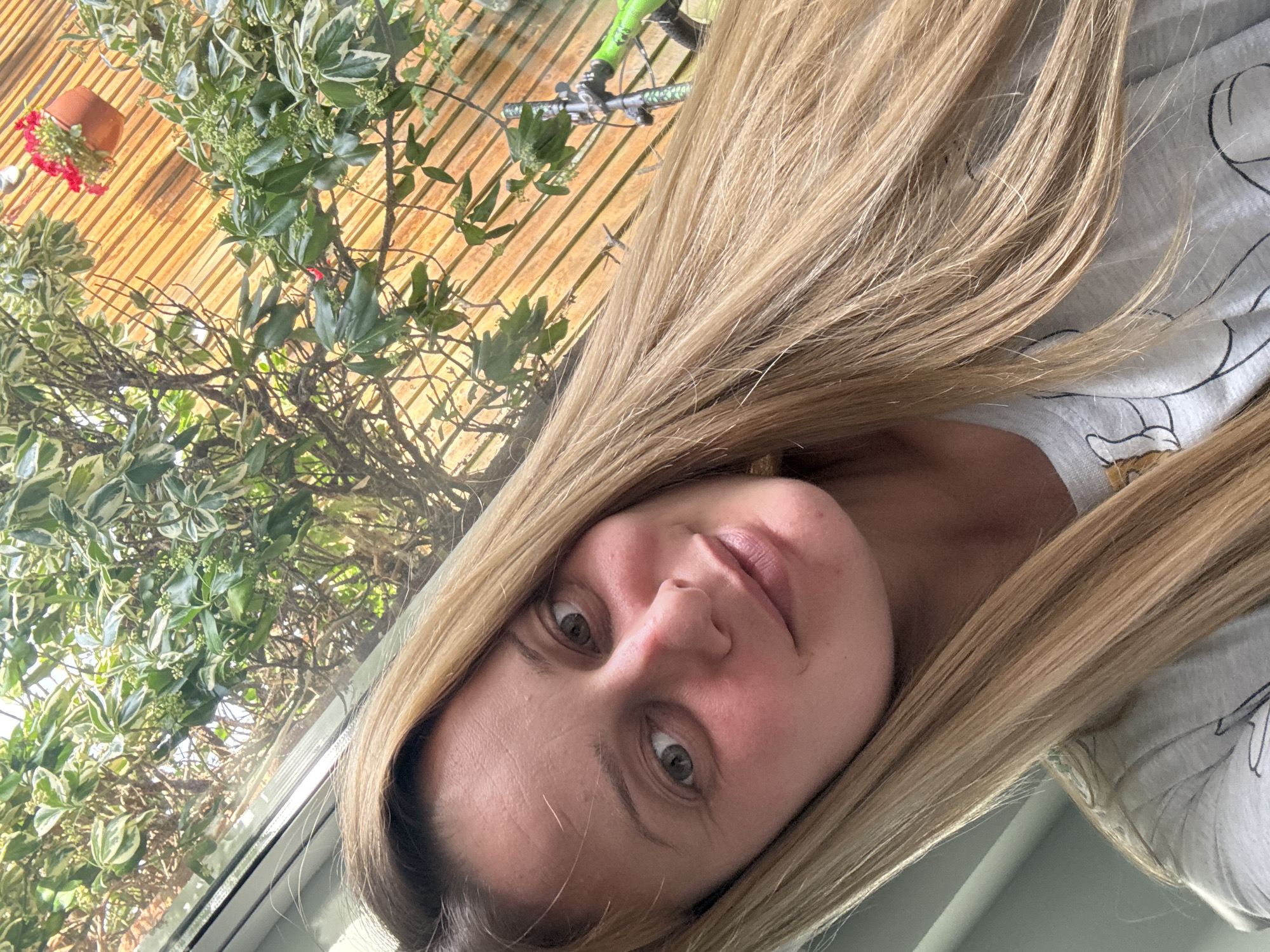 A woman with long blonde hair is seen at a close-up angle indoors, possibly in Hazlemere, United Kingdom, given the prompt. She is wearing a light-colored t-shirt with cartoon designs. Behind her, visible through a window or door, is a wooden structure, possibly a fence or shed, with plants growing against it. A small terracotta pot with red flowers is also visible. A bicycle can be seen in the background on top of the wooden structure.