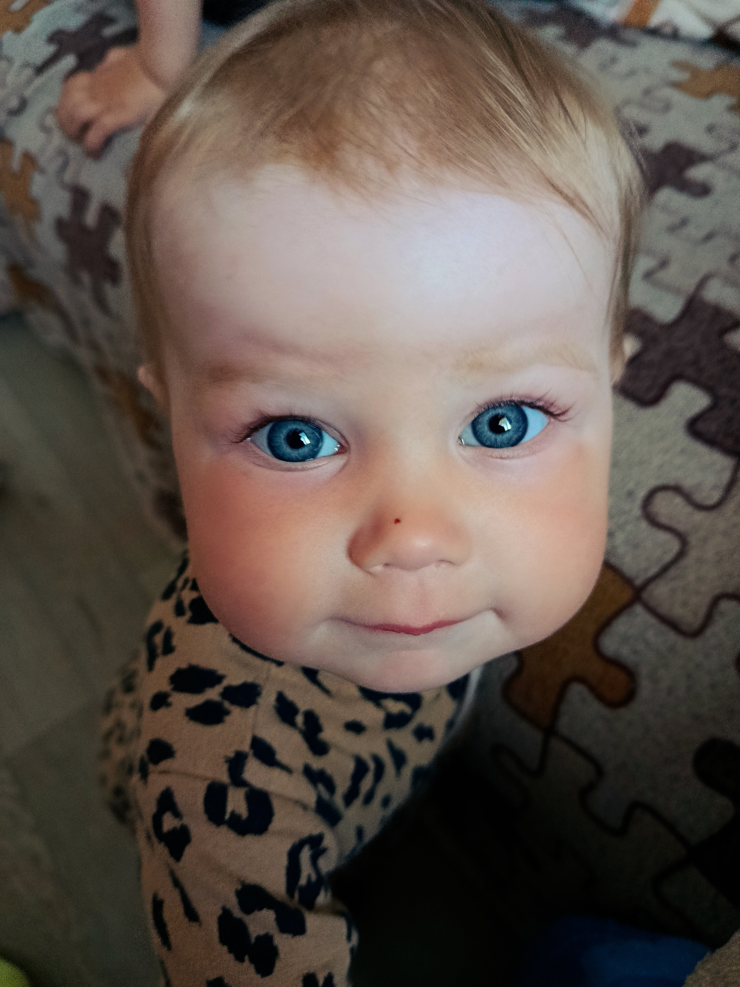 This is a close-up, slightly high-angle shot of a baby's face, filling most of the frame. The baby has fair skin, light brown hair, and striking blue eyes that are looking directly at the camera. There is a small, reddish mark on the bridge of the baby's nose. The baby is wearing a tan shirt with a black leopard print pattern. The background appears to be a textured surface, possibly a blanket or upholstery, with a puzzle piece pattern in shades of brown and gray. A small part of another person's leg and foot is visible in the upper left corner. The image appears to be taken indoors, in a residential setting. The location of Фоки, Russia is not discernible from the image content.