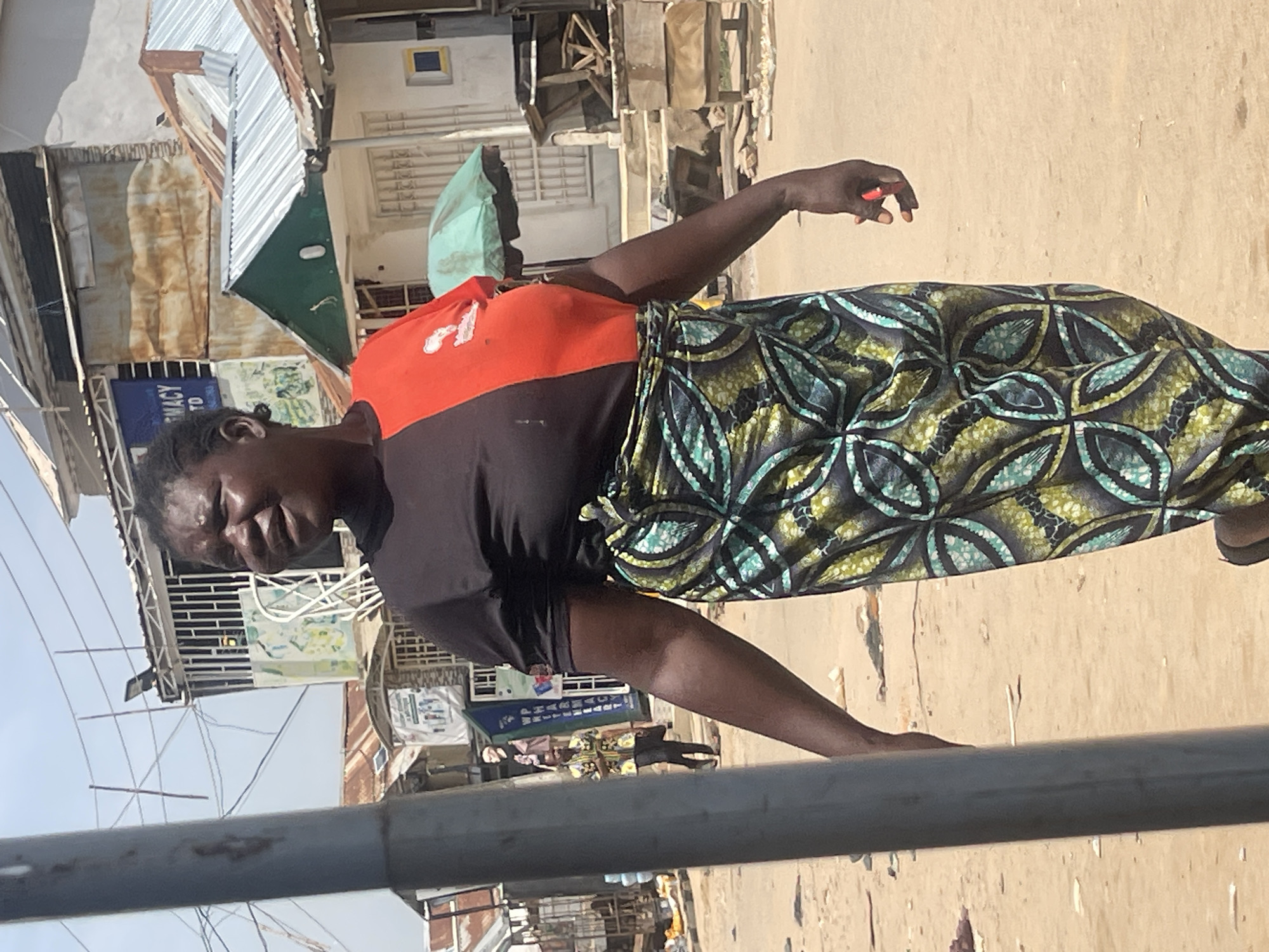 A woman in an orange and black top and a patterned skirt stands in what appears to be a market or bustling street scene. The setting is outdoors with several buildings and structures in the background, some with corrugated metal roofs and exposed construction. There are signs visible, including one that reads "PHARMACY TO". The ground is dusty or dirt. The woman's expression suggests she may be speaking or reacting to something, perhaps with a hint of concern or exertion. Her right hand is slightly raised, with what looks like nail polish on one finger. The overall lighting suggests it is daytime, and the environment appears to be an urban or semi-urban area in Nigeria, likely Abuja, given the provided location context. The image is captured in a portrait orientation.