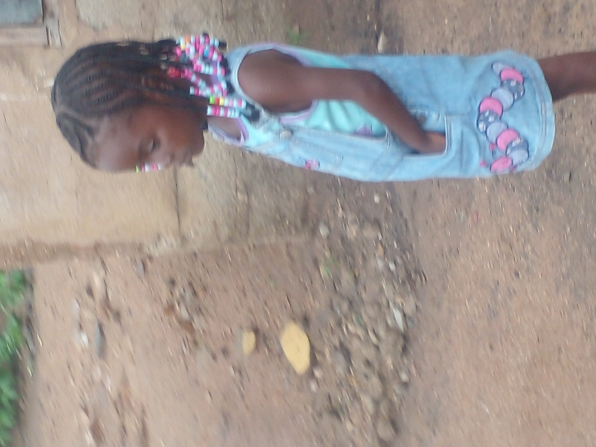 In the city of Unknown, Unknown, a young Black girl stands outdoors, her body angled away from the camera and turned slightly to the left. She has braided dark hair, adorned with colorful beads. She is wearing a light blue denim overall dress over a light blue top. Her left hand is tucked into the pocket of her dress. The ground around her is composed of dirt and small stones, with some scattered vegetation in the background. A light-colored wall is visible behind her. The image appears to be taken during the daytime.