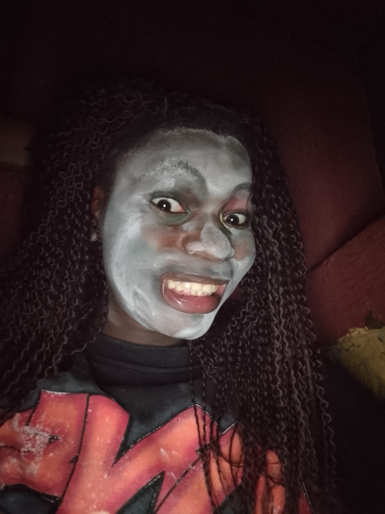 The image is a close-up, slightly off-center portrait of a person in an indoor setting in Onitsha, Nigeria. The individual, who has dark skin and long, dark, curly braided hair, is looking directly at the camera. Their face is covered in a light gray or white substance, resembling a face mask or theatrical paint, with subtle reddish smudges around the eyes and nose areas.
The person displays an exaggerated expression, featuring wide-open eyes and a broad, open-mouthed smile that reveals their teeth and tongue. This creates a playful, wild, or potentially "spooky" demeanor. They are wearing a dark, possibly black, long-sleeved shirt or sweater. On the front of the shirt, a large, prominent graphic in distressed orange and red is visible, identifiable as the logo for the band KISS. White, textured details appear on the red lettering.
The background is dimly lit and out of focus, showing a dark red or maroon upholstered surface, likely part of a sofa or chair, on the right side. A small, lighter, textured object, possibly yellow or gold, is partially visible in the bottom right corner. The low lighting suggests the image was taken indoors, possibly at night or in a dimly lit room.