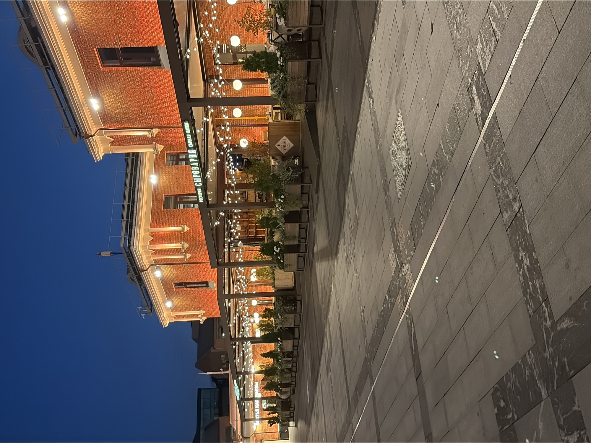 The image captures an exterior view of the restaurant "Syrovar'ya" (Сыроварня) in Moscow, Russia, at twilight. The restaurant is housed in a brick building with illuminated windows and decorative architectural elements. An outdoor seating area is set up with a canopy strung with numerous string lights, creating a festive ambiance. Planters filled with greenery and small trees are arranged along the edge of the seating area, separating it from a paved walkway. The walkway itself is constructed with large, dark gray stone pavers, interspersed with decorative bands of lighter stone and a metallic strip running down its center. In the distance, other buildings are visible, including a modern, darker structure on the left. The sky above is a deep, clear blue, indicative of early evening. There are no people visible in the frame. The scene conveys a sense of an urban evening setting, with the restaurant's lighting providing warmth against the darkening sky.