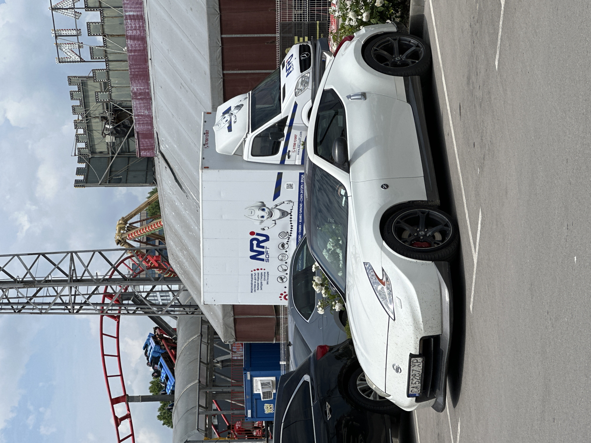 The image shows a white Nissan 370Z parked in a lot next to a white van and a dark car. The van has "NRI Soft" and a phone number on its side. In the background, there is a roller coaster and other amusement park structures. The license plate of the car is CA5287XP, which indicates that the car is registered in Bulgaria. The van also contains text in Bulgarian. Given the presence of the roller coaster and the Bulgarian text, the location is likely an amusement park in Varna, Bulgaria.