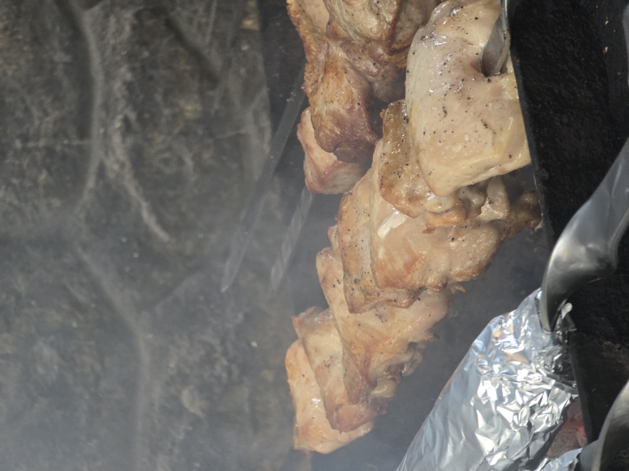 The image shows a close-up view of meat skewers cooking on a grill, possibly in Pidhorodne, Ukraine. The meat, appearing to be chicken or pork, is browned and seasoned with pepper. It is threaded onto metal skewers and arranged horizontally on the grill. Smoke is visible, indicating the cooking process. In the foreground, a skewer is partially wrapped in aluminum foil, and a metal utensil is also visible. The background consists of a rough, textured stone surface, which could be part of a grill enclosure or a decorative element. The scene is dimly lit, with the focus on the food.