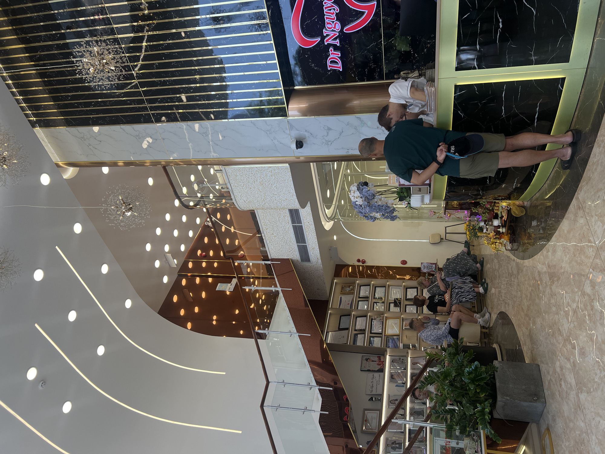 The image shows the interior of a building. The building is well-lit, featuring a modern design with white walls, decorative lighting, and marble-like surfaces. A man in a green shirt is standing at a reception desk, interacting with a person behind it. Seated in a waiting area are several people. There are framed pictures or documents on the wall. A staircase with glass railings is visible.
Based on the “Dr. Nguyen” sign, the location is Nha Trang, Vietnam.