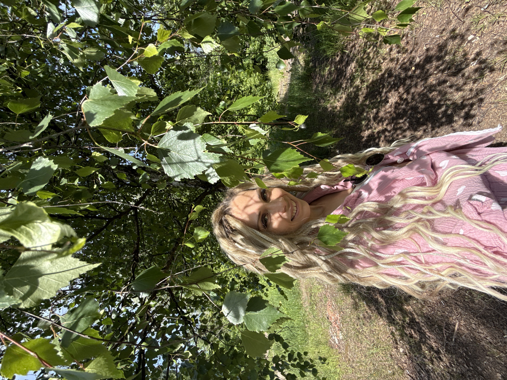 The image displays a single female subject positioned outdoors in a natural, wooded environment, likely a park or forest edge, within the city of Rusanovo, Russia. The woman, shown from the chest up, has long, wavy blonde hair with integrated lighter-colored dreadlocks or braids. She is smiling directly at the camera, with a portion of her face obscured by shadows cast by overhead leaves. She is wearing a light pink garment, possibly a loose top or dress, with a crinkled texture and faint lighter patterns. Dark-lensed sunglasses are visible, hooked onto the front of her attire.
The foreground and upper portion of the image are dominated by numerous vibrant green leaves and slender reddish-brown branches of a deciduous tree, consistent with a birch, which partially frame the woman. Bright sunlight illuminates the scene from above, creating distinct dappled shadows on the ground behind and to the side of the woman, and highlighting various leaves. The ground consists of patches of green grass interspersed with areas of exposed soil and dry leaves, suggesting an unpaved path or natural terrain. In the background, denser green foliage indicates further trees and undergrowth, with glimpses of a clear blue sky through the upper canopy.