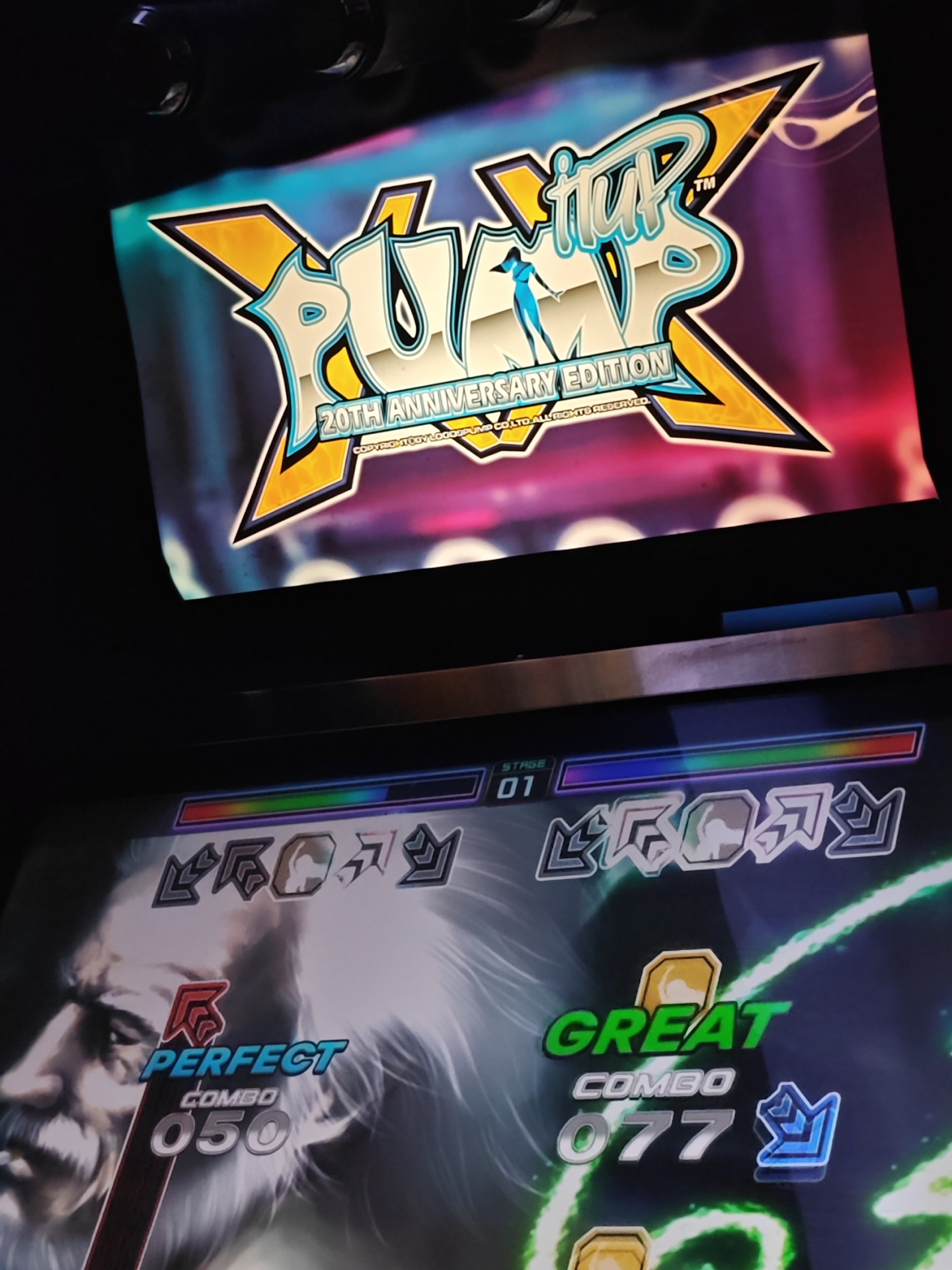 The image depicts the screen of a "Pump It Up XX 20th Anniversary Edition" arcade game. The top screen displays the game's logo, while the bottom screen shows the gameplay interface. On the gameplay screen, the left side reads "Perfect Combo 050" with an image of a man with white hair and a mustache, while the right side reads "Great Combo 077." The stage number "01" is displayed on the top of the bottom screen. The scene captures the experience of playing the Pump It Up arcade game. There is no information about the location in the city of Barcelona, Spain, nor essential details about people, subjects, objects, actions, interactions, and the environment.