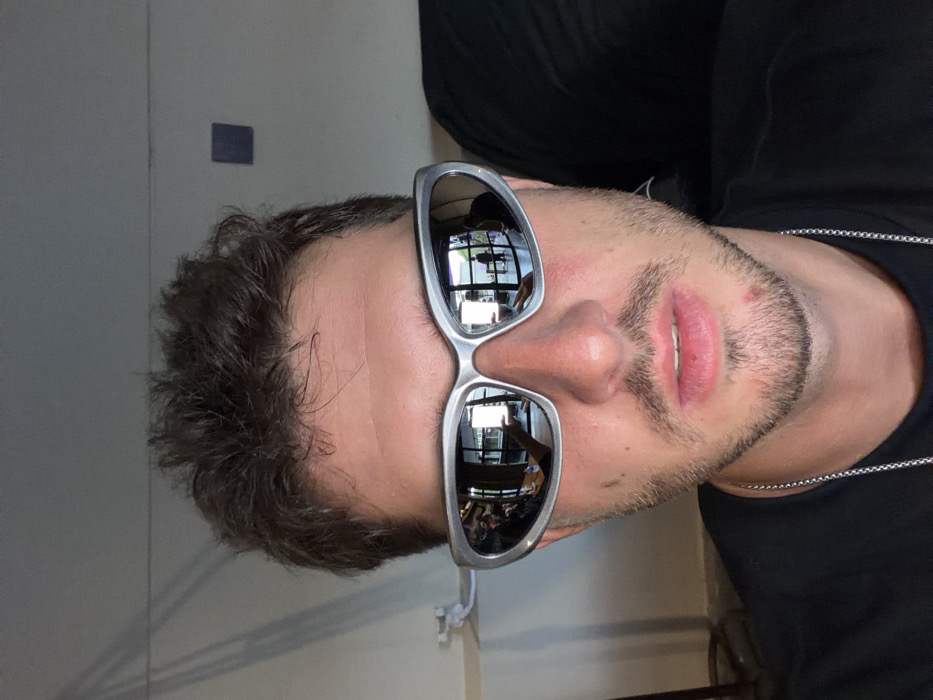 A close-up, head-and-shoulders portrait features a male individual with short, dark brown hair and light facial stubble. The subject wears silver-framed, reflective, wraparound sunglasses, a black t-shirt, and a silver-colored chain. A minor blemish is visible on the subject's chin and lower lip.
The reflections within the sunglasses reveal an indoor environment located in Tumbak Bayuh, Indonesia. The reflections show a spacious area characterized by large, dark-framed windows or glass doors. In the left lens, a person's arm and hand are visible, potentially holding an object, alongside a wooden surface and a blurred individual. The right lens reflects a bright rectangular object, an additional standing figure, and further window elements.
The background directly behind the subject consists of a plain, light-colored wall. Subtle vertical shadows, consistent with blinds, are visible on the upper left wall section. A small, dark blue square object is affixed to the upper right wall. A white fixture is partially visible on the left side, near the subject's left shoulder. An out-of-focus dark object occupies the right edge of the frame.