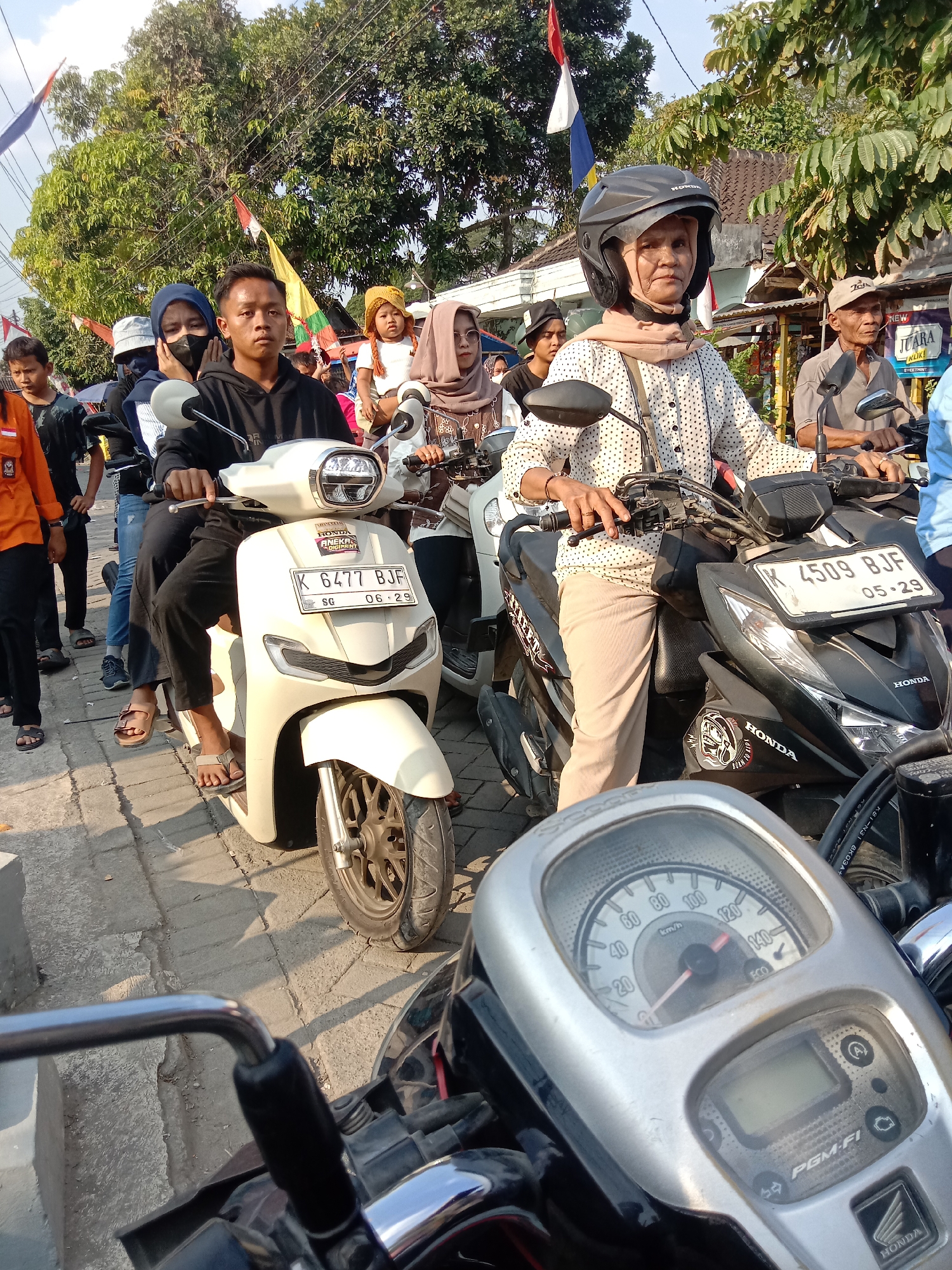 The image depicts a street scene in Wirosari, Indonesia, likely during a daytime event. A procession of people on motorcycles is evident, with flags and decorations suggesting a local celebration or parade. In the foreground, a white scooter with license plate K 6477 BJF is prominently featured, driven by a young man wearing a black hoodie. Behind him, several individuals are seated, including a woman in a blue hijab and another wearing a black mask and a blue headscarf. To the right, a black Honda scooter is driven by an older woman wearing a helmet and a light-colored patterned shirt, with an elderly man in a brown shirt and cap riding behind her. Further back, other participants are visible, including children. The environment includes paved sidewalks, trees, and buildings lining the street, with overhead wires suggesting a residential or commercial area. The overall atmosphere appears to be one of community engagement and public gathering.