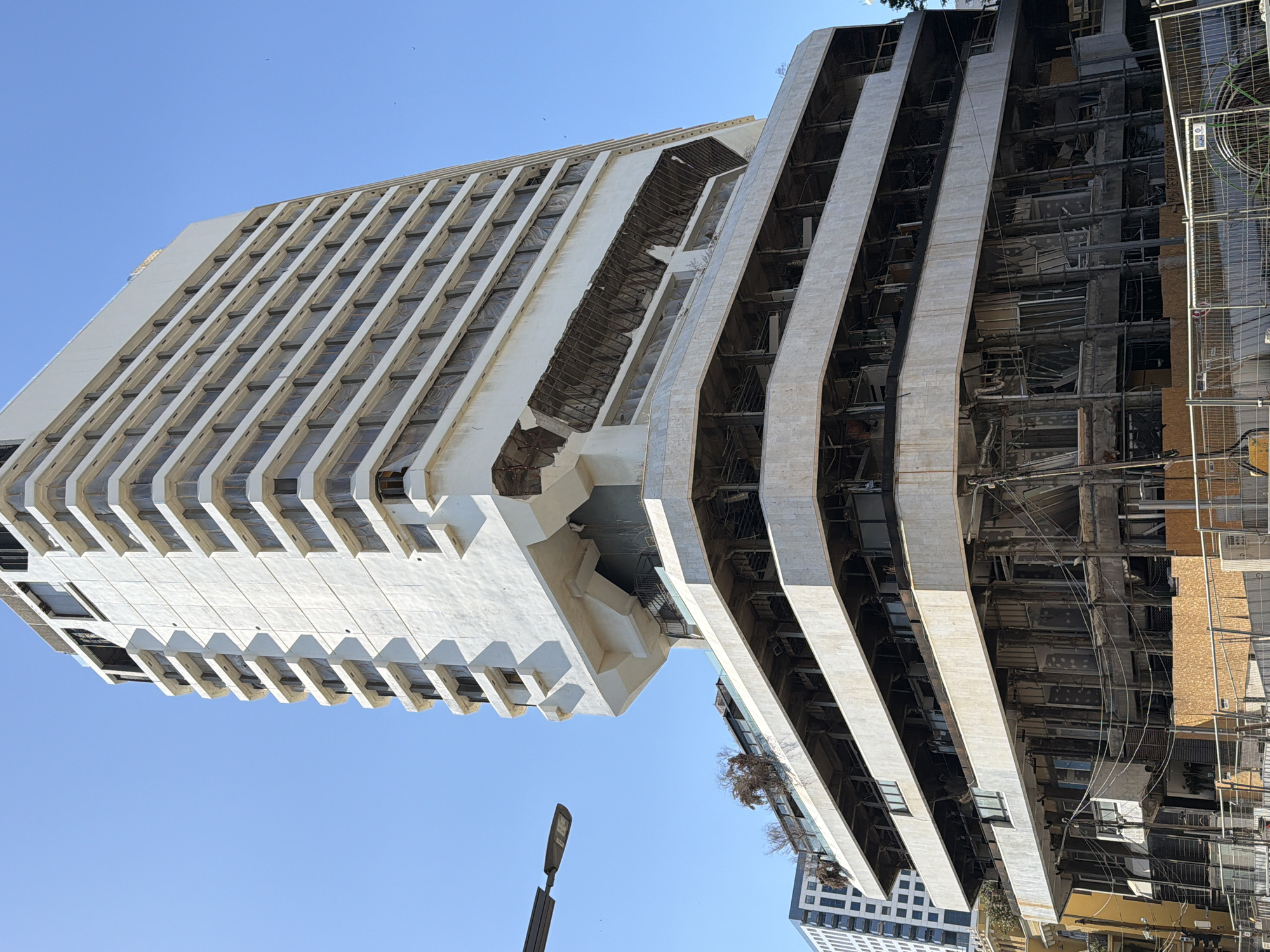 The image shows a tall building in Tel-Aviv, Israel, in a state of disrepair. One side of the building is mostly intact, with white concrete and rows of rectangular windows. The other side is heavily damaged, revealing exposed internal structures and debris. A light pole is visible in the lower-left corner of the image. A chain-link fence is visible along the bottom right edge. The sky is clear and blue. Another, smaller building is visible at the bottom right corner. The building is undergoing deconstruction/reconstruction.