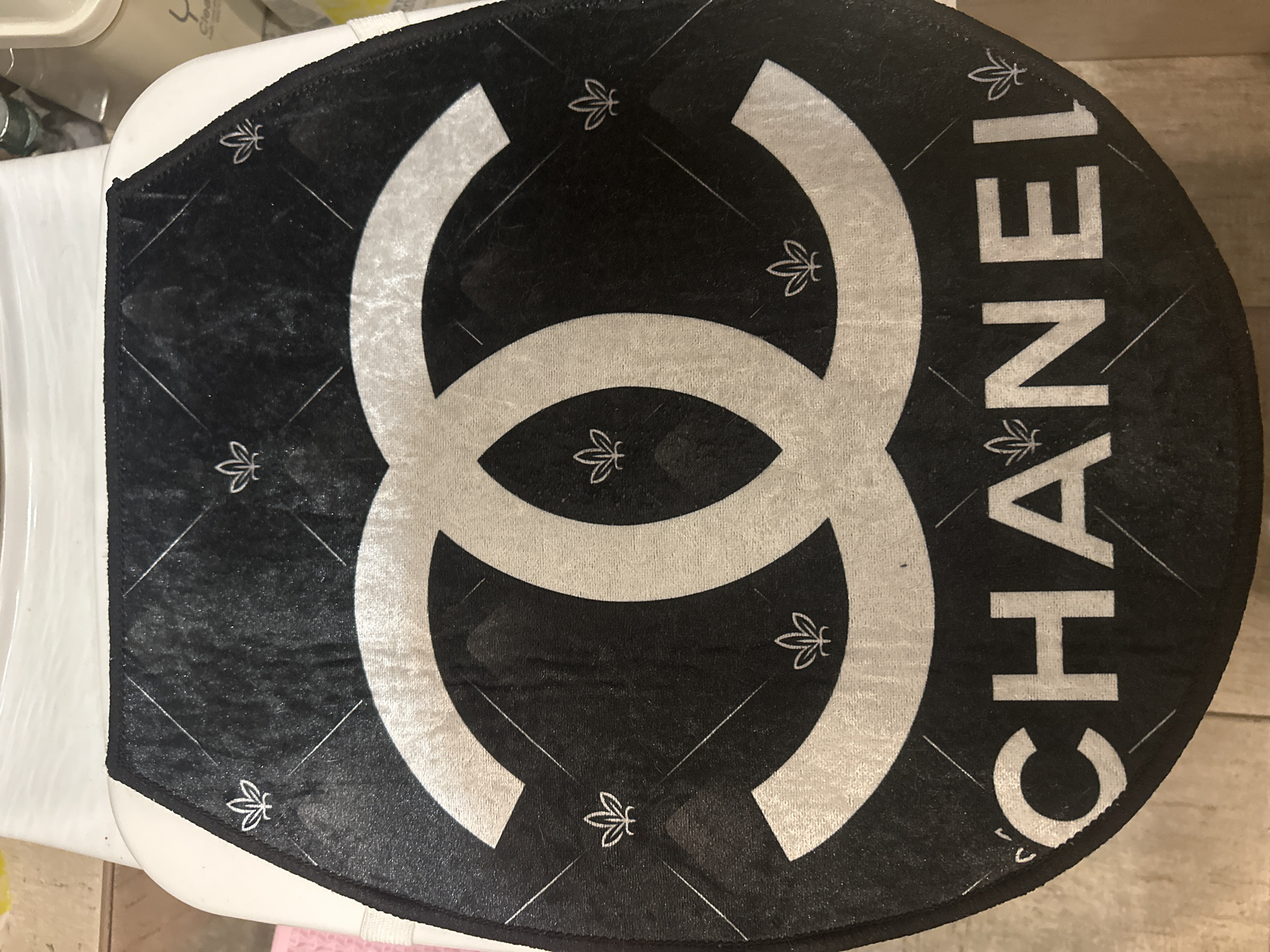 A black toilet seat cover with a white Chanel logo and the word "CHANEL" is displayed on a toilet. The seat cover has a diamond-shaped quilted pattern with small leaf-like symbols in each diamond. The Chanel logo, consisting of two interlocking C's, is prominently featured in the center of the seat cover. The word "CHANEL" is written in white capital letters at the bottom of the seat cover. The background is blurred and appears to be a tiled floor. This image was taken inside a restroom in Kyiv, Ukraine.