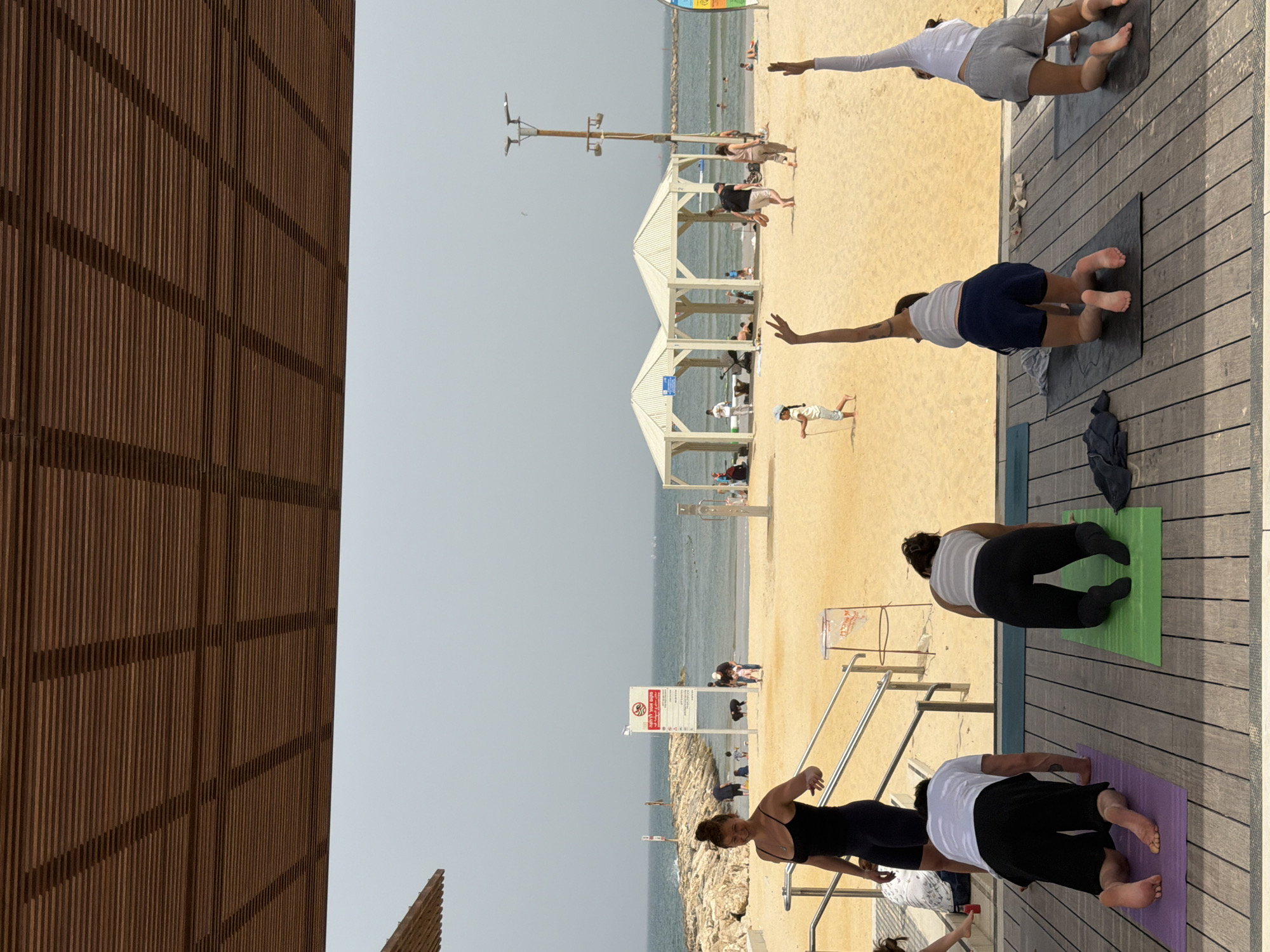 The image shows a group of people doing yoga on a wooden deck at a beach in Tel Aviv, Occupied Palestine.
There are several people in yoga poses on yoga mats. The yoga mats are positioned on the wooden deck which is adjacent to a sandy beach. In the background, there are canopies and the ocean, with some individuals on the beach. A wooden structure is visible on the left side of the frame. A sign is visible near a set of stairs leading from the wooden deck to the beach.