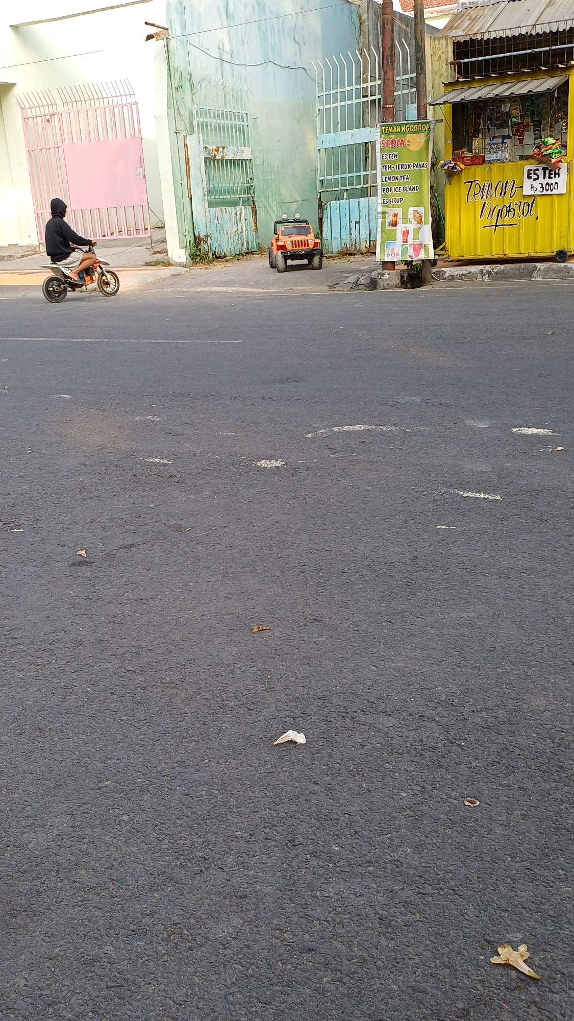 In Klaten, Indonesia, a person wearing a black hoodie and shorts rides a small dirt bike on a paved road. To the left of the road, there is a pale pink gate. Behind the gate, a building with a light green facade and white bars is visible. To the right of the building, there is a yellow food stall with a sign that reads "TEMAN NGOBROL" and lists "ES TEH," "ES TEH JERUK PANAS," "LEMON TEA," "POPCICE BLAND," and "ES SIRUP." Next to the stall, an orange toy jeep is parked on the sidewalk. The sky is overcast.