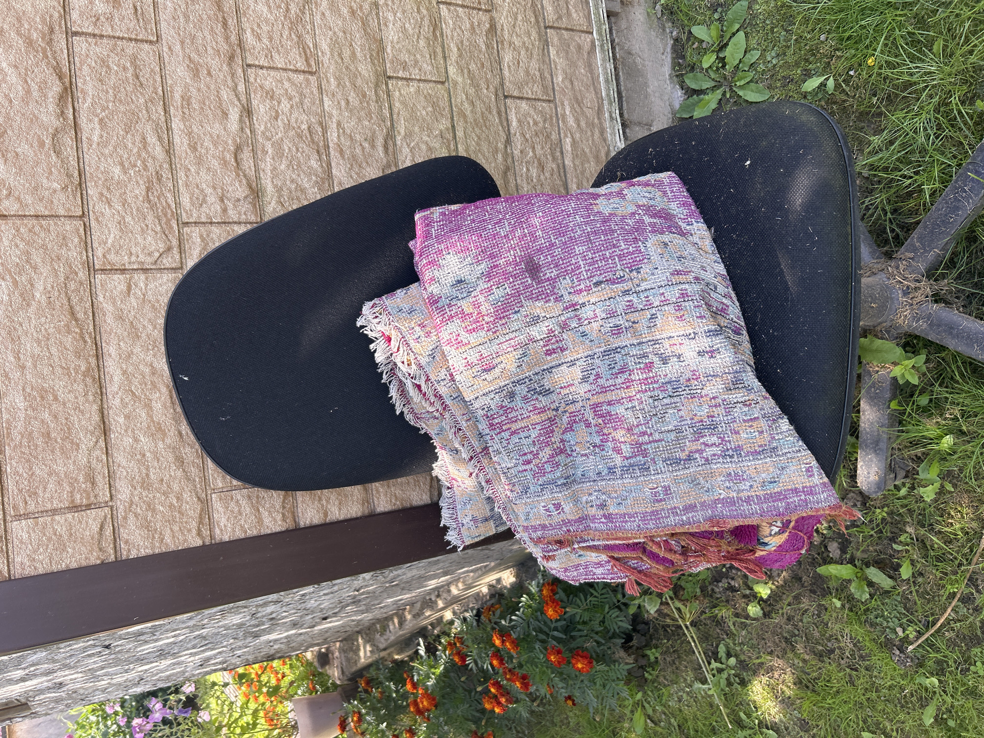 The image depicts an office chair with a folded colorful blanket placed on its seat, situated outdoors in front of a brick wall. The environment appears to be a yard or garden, with green grass and some flowering plants visible to the left of the chair. The brick wall shows a tan, textured surface with visible mortar lines. The chair itself is black, with visible signs of wear and dirt. The blanket is intricately patterned with shades of pink, purple, blue, and yellow, and features fringed edges. To the left of the chair, a row of vibrant orange flowers and some taller purple flowers are in bloom. The overall scene suggests an outdoor setting, possibly in a residential area. The location is identified as Kalitsino, Russia.