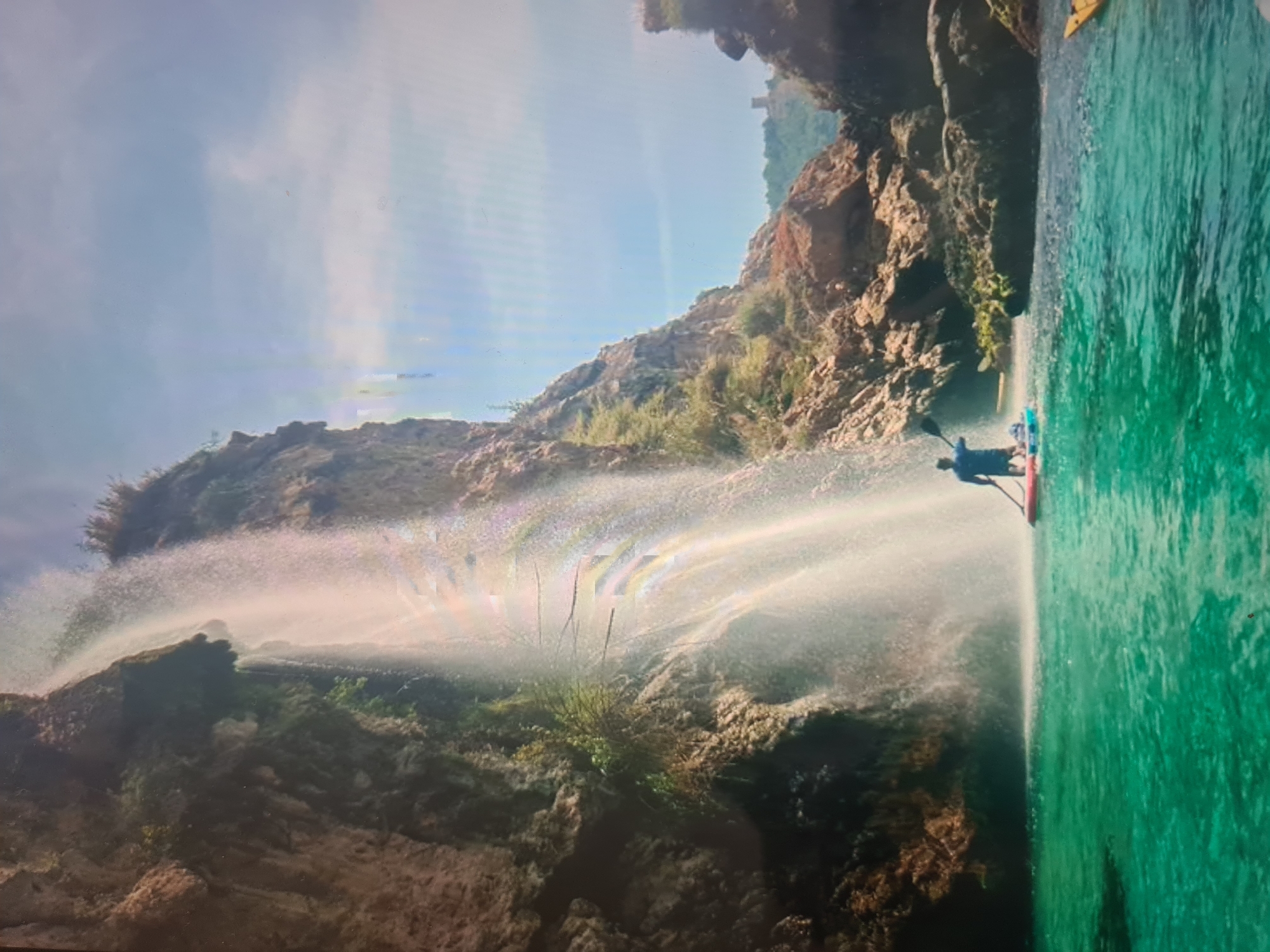 A vibrant outdoor scene captures a person paddleboarding near a powerful waterfall. The image, seemingly taken on a bright, sunny day, showcases a natural landscape characterized by towering rocky cliffs and clear, emerald-green water.
In the foreground, a person is seen from behind, standing on a red and white paddleboard and holding a paddle. They are situated in the calm, transparent water, close to the tumultuous spray of the waterfall. The waterfall itself cascades down a rugged, moss-covered cliff face on the left, creating a broad sheet of mist and spray that brilliantly catches the sunlight, casting bright reflections.
The rocky cliffs, covered in patches of green shrubs and sparse vegetation, dominate the left and background of the scene, rising sharply towards a bright blue sky with scattered white clouds. A portion of a yellow kayak or small boat is faintly visible further out in the water on the right, suggesting more activity. The overall impression is one of adventure and natural beauty in a serene, untamed environment. There is no visible text in the image.